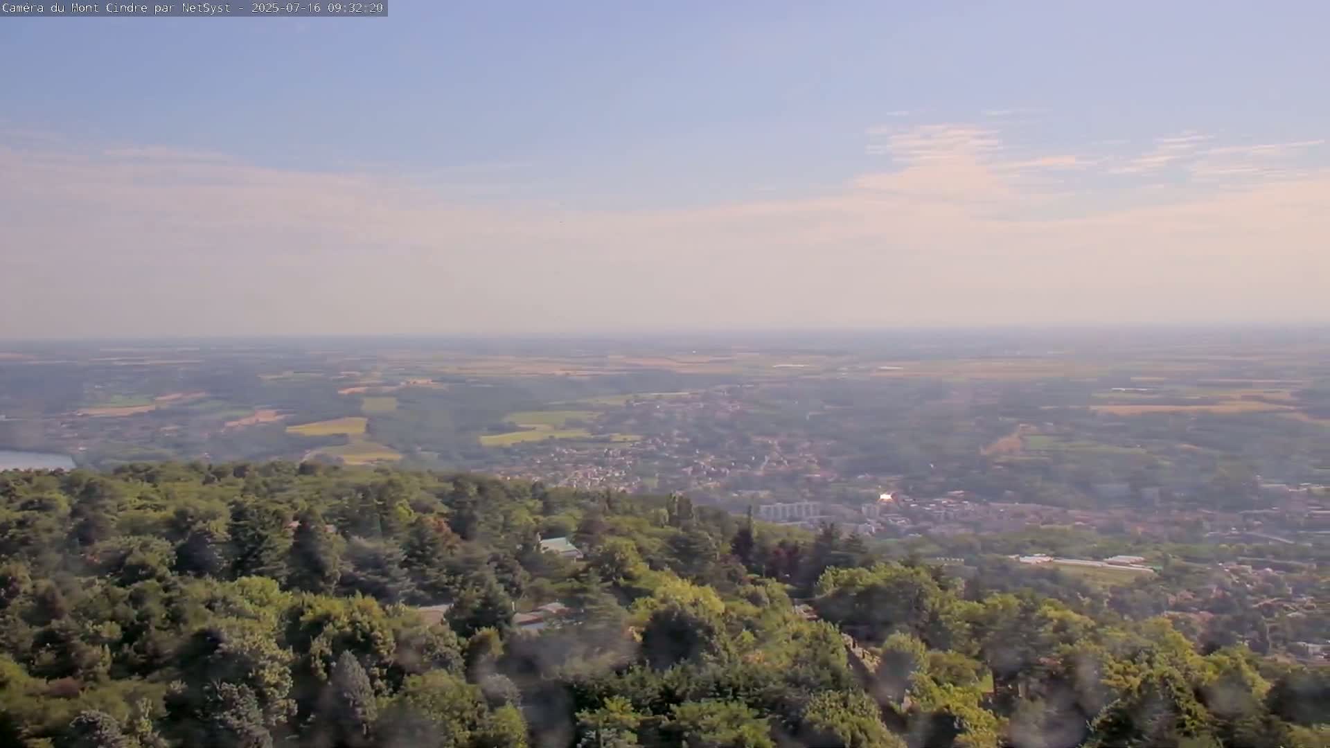 A hazy, slightly cloudy day reveals a panoramic view of a town nestled in a valley, surrounded by lush green forests, from a high vantage point.