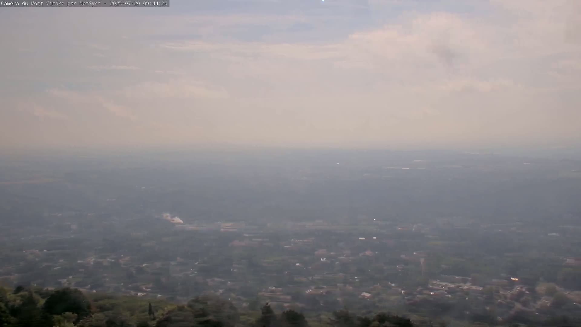 A hazy, partly cloudy day reveals a sprawling town or city viewed from a high vantage point, with trees and hills in the foreground.