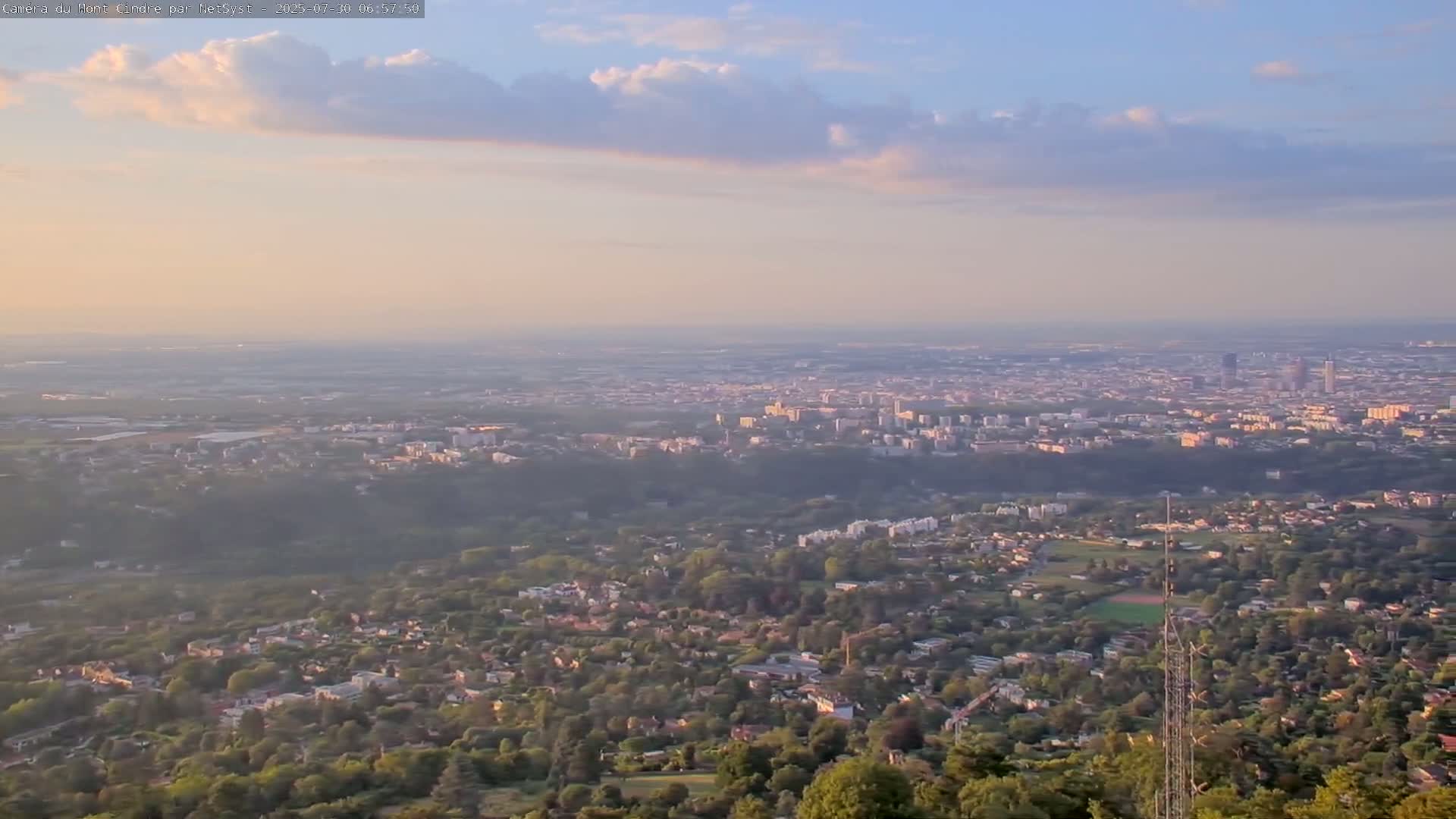 A hazy, partly cloudy day reveals a sprawling town or city viewed from a high vantage point, with trees and hills in the foreground.