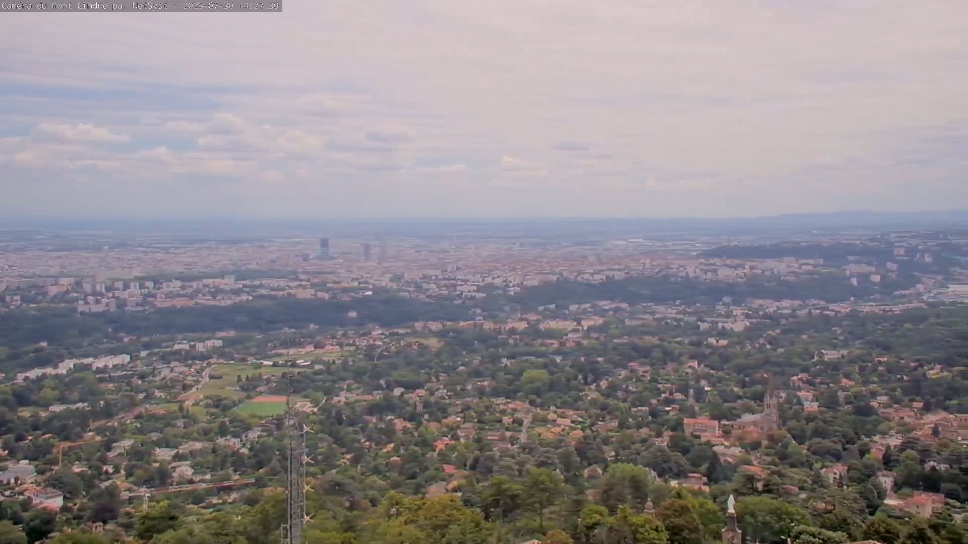 A hazy, partly cloudy day reveals a sprawling town or city viewed from a high vantage point, with trees and hills in the foreground.