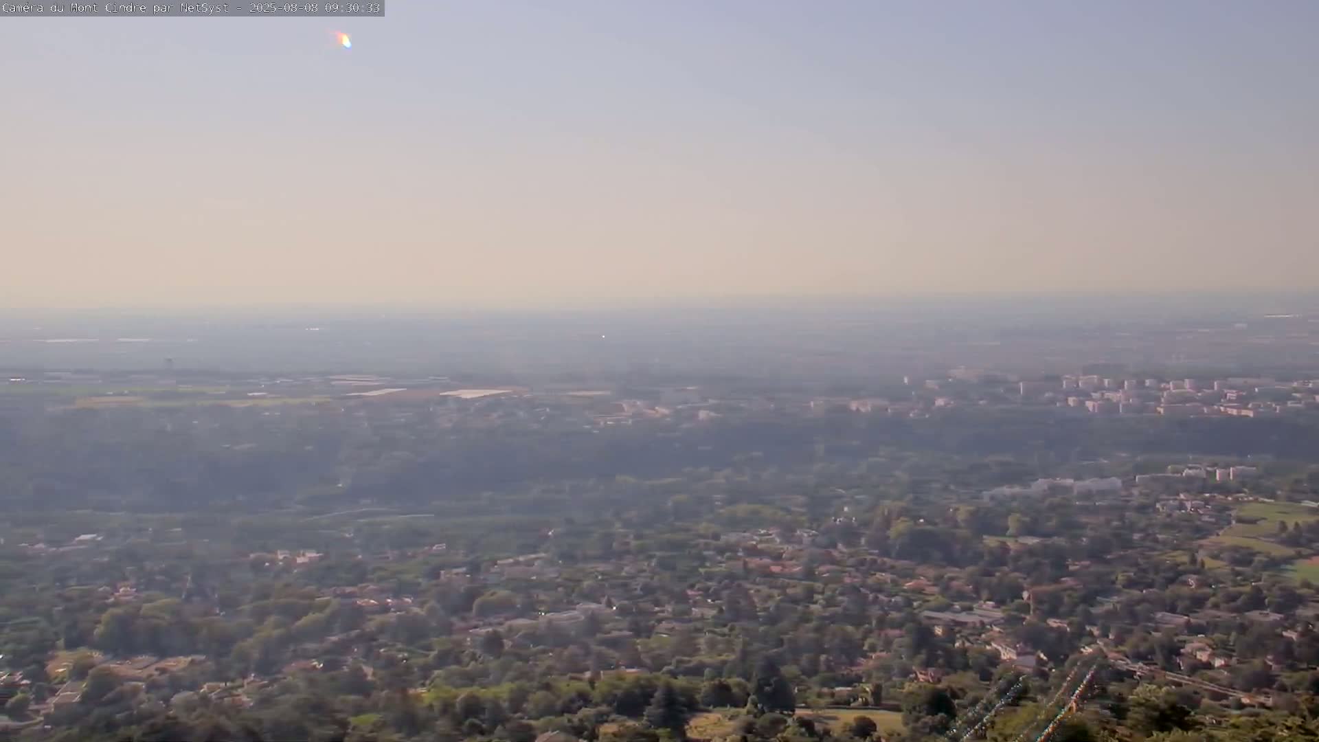 A hazy, long-distance view shows a sprawling suburban area with green trees and buildings extending to a distant horizon under a light-blue sky.