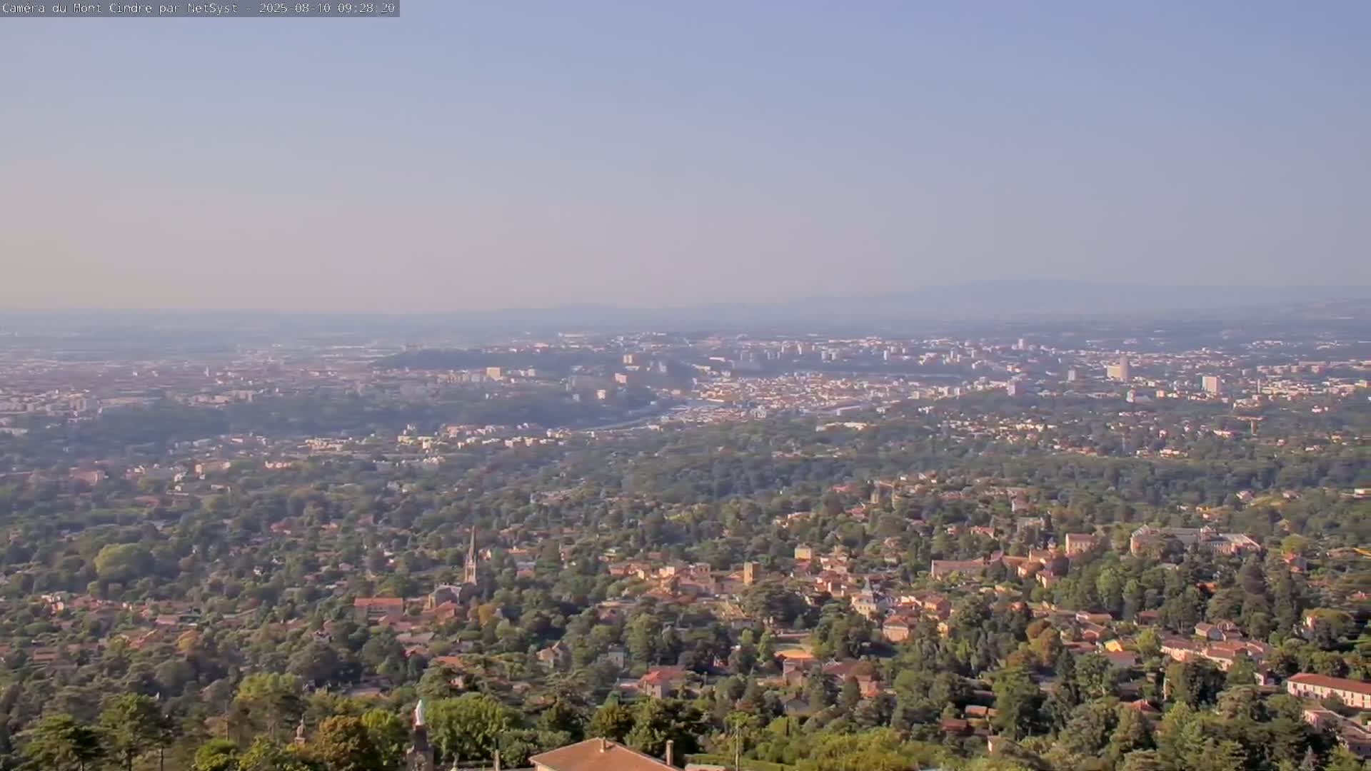 A hazy, long-range view shows a sprawling city nestled amongst lush green hills and forests, extending to a distant horizon.