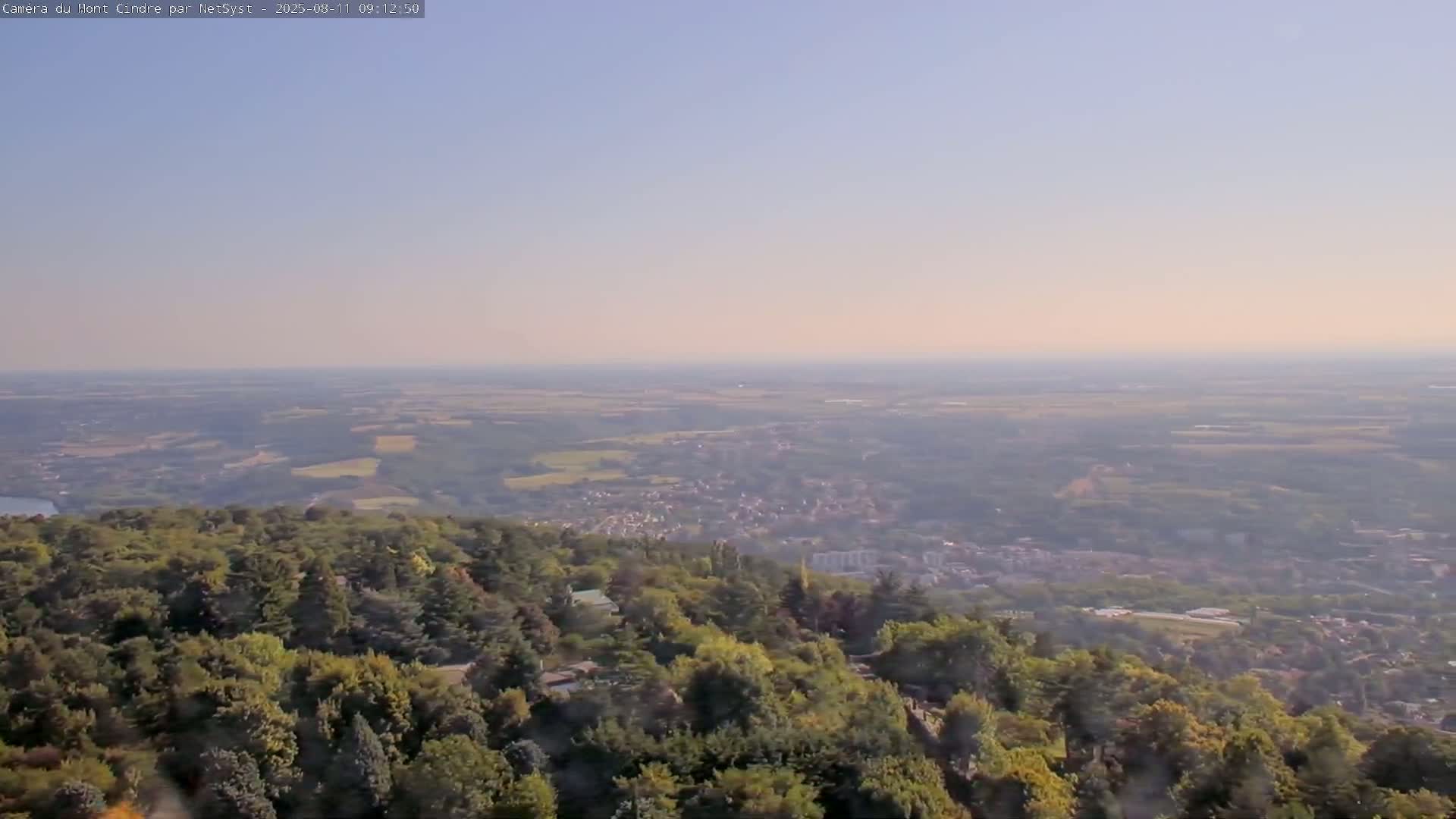 A hazy, sunny day reveals a vast landscape of rolling hills, farmland, and a town nestled amongst a lush, green forest from an elevated viewpoint.