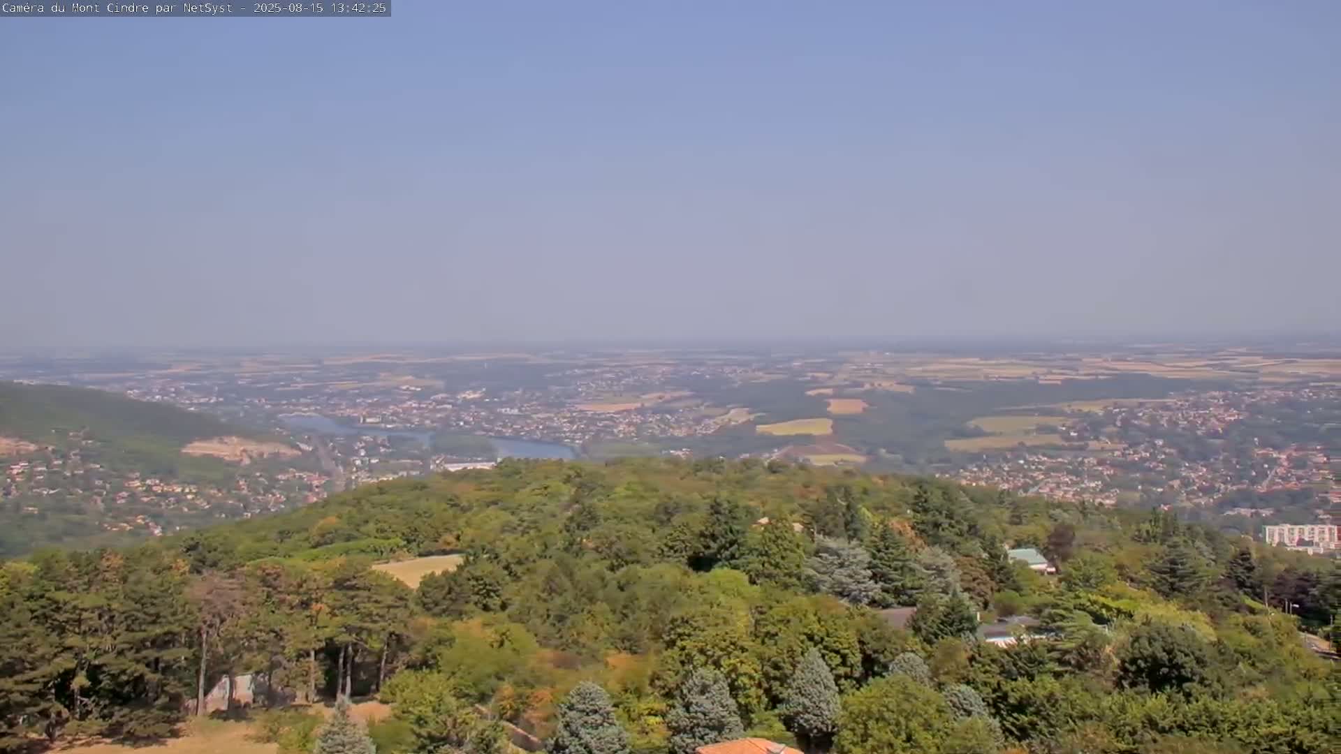 A hazy, sunny day reveals a vast landscape of rolling hills, farmland, and a town nestled amongst a lush, green forest from an elevated viewpoint.