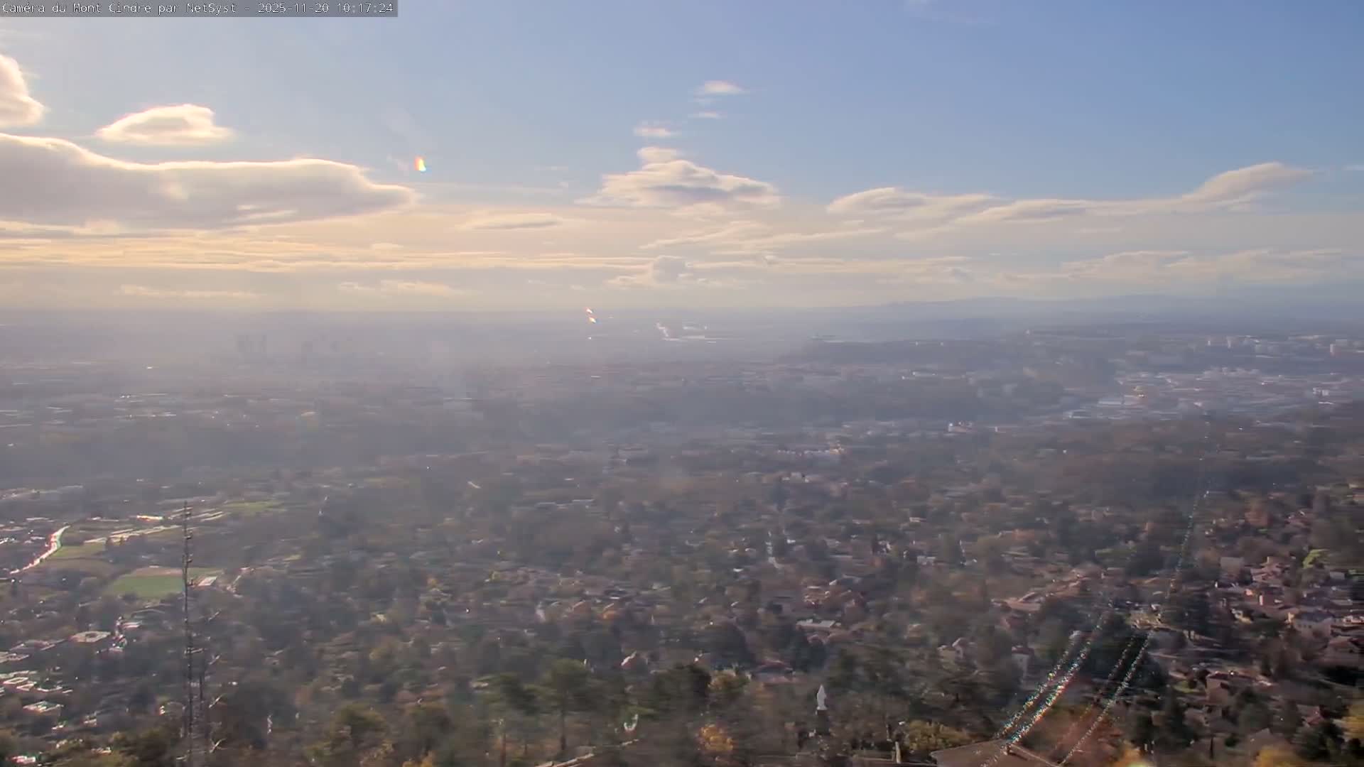 A sweeping high-angle view reveals a sprawling urban landscape dotted with trees, partially obscured by haze under a brightly lit, partly cloudy blue sky.