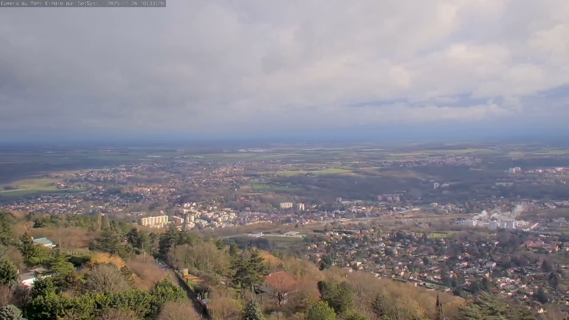 A wide, high-angle view captures a sprawling town nestled in a valley, surrounded by a mix of wooded hills and distant cultivated plains, all under a partly cloudy sky.