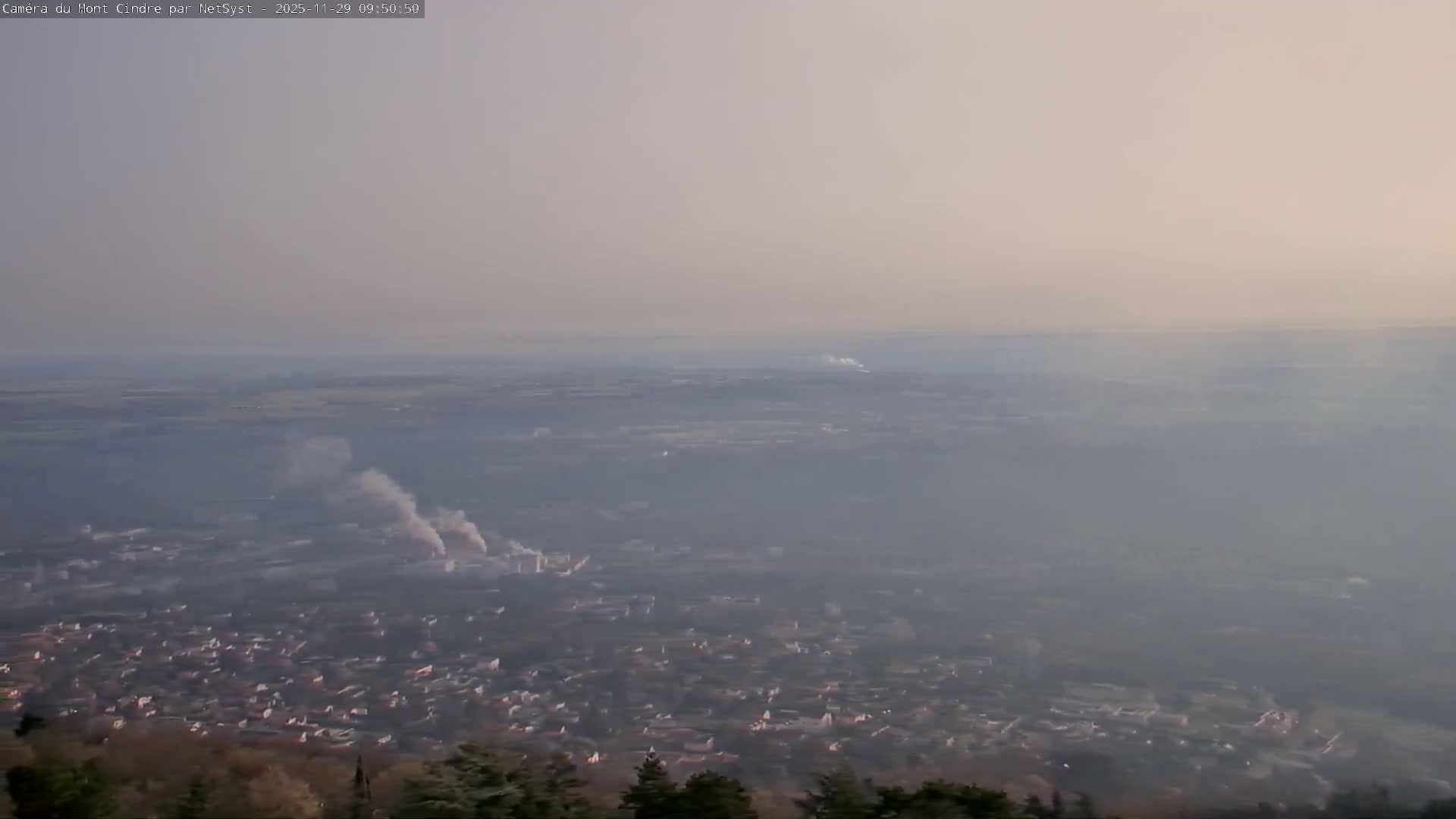 An expansive, hazy view under an overcast sky reveals a city below with multiple plumes of smoke rising from industrial areas, and indistinct distant landscapes.