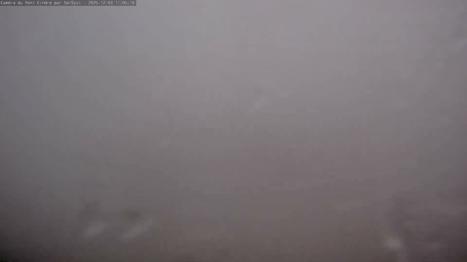 The image shows an outdoor scene completely obscured by dense fog or clouds, resulting in extremely low visibility with only faint, blurry dark shapes dimly visible towards the bottom.