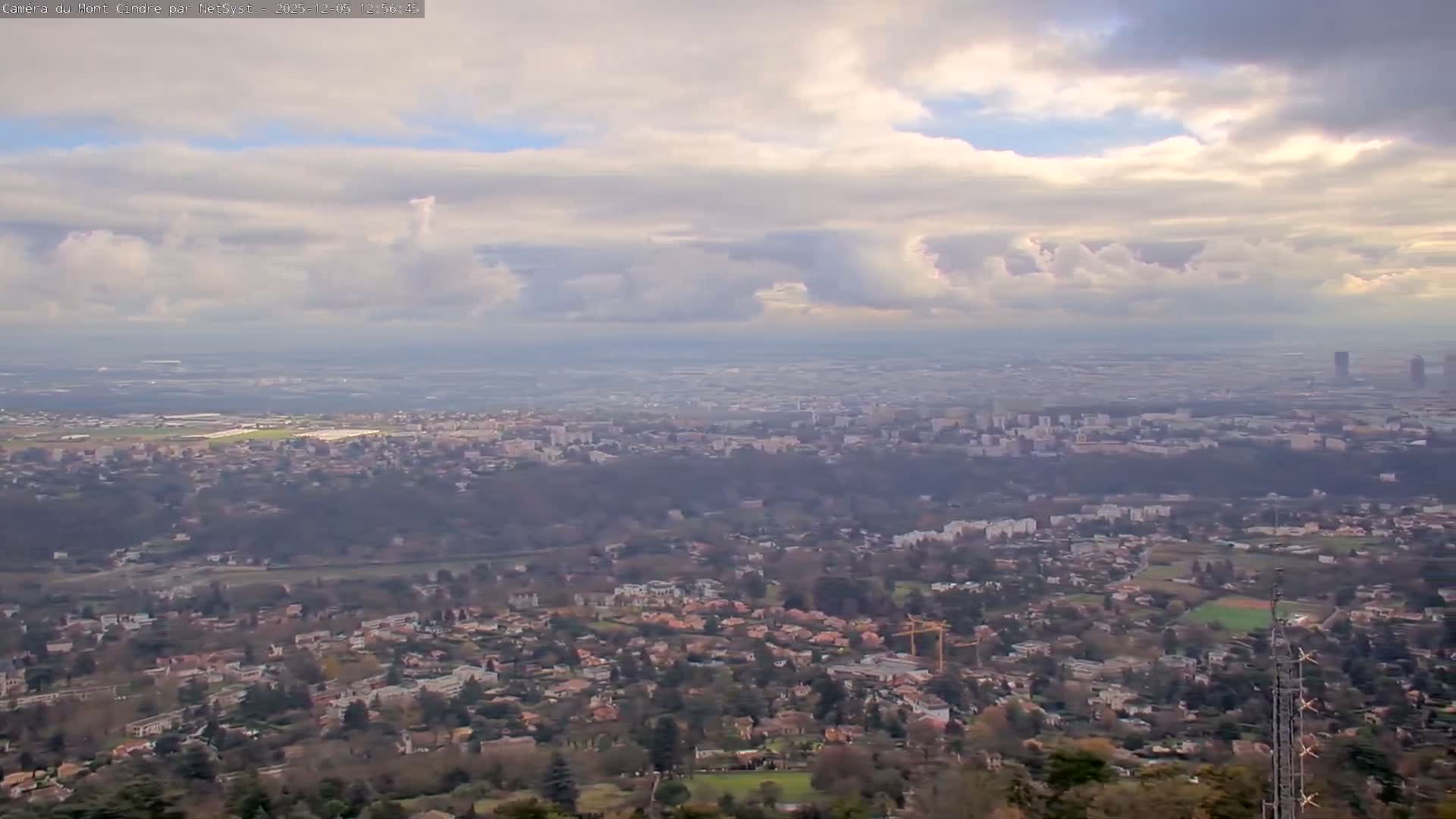 An expansive aerial view captures a sprawling city under a partly cloudy to overcast sky, with residential areas and trees in the foreground transitioning to dense urban development extending to a hazy horizon.
