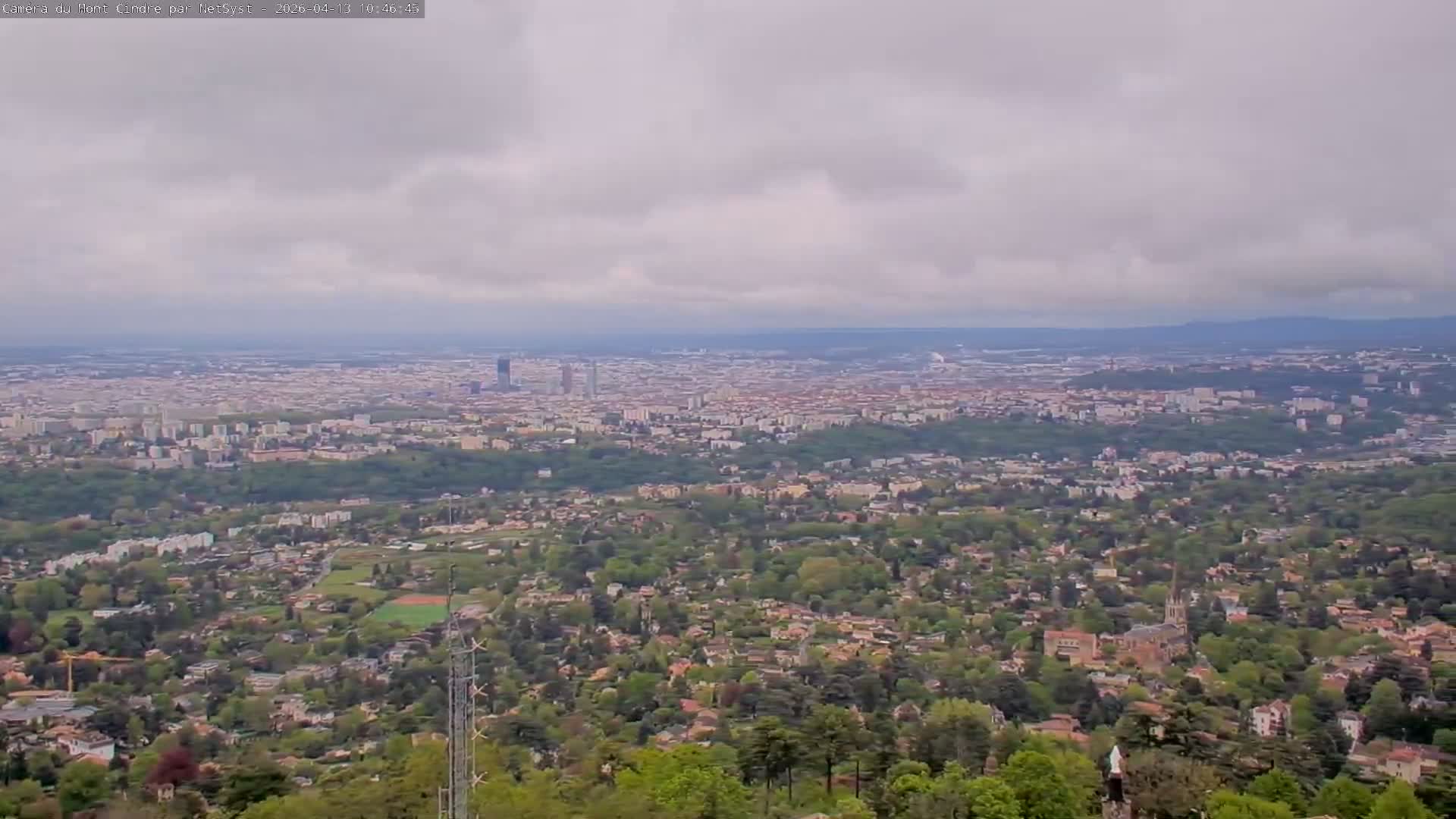 An expansive aerial view captures a sprawling city under a partly cloudy to overcast sky, with residential areas and trees in the foreground transitioning to dense urban development extending to a hazy horizon.