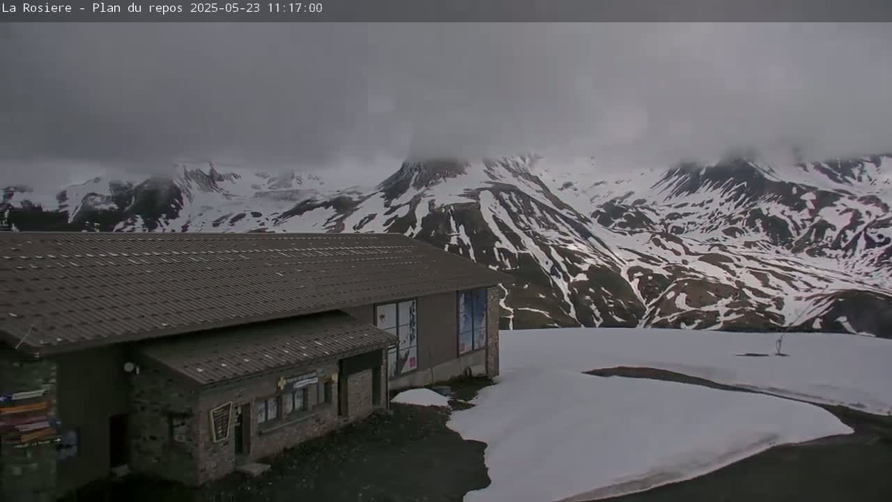 A low stone building with a dark gray roof sits on a snow-covered mountainside under a cloudy sky.