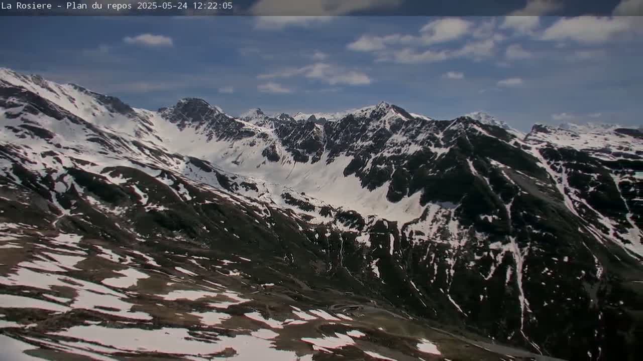 A partly cloudy sky overlooks a snow-dusted mountain range with dark, rocky slopes.