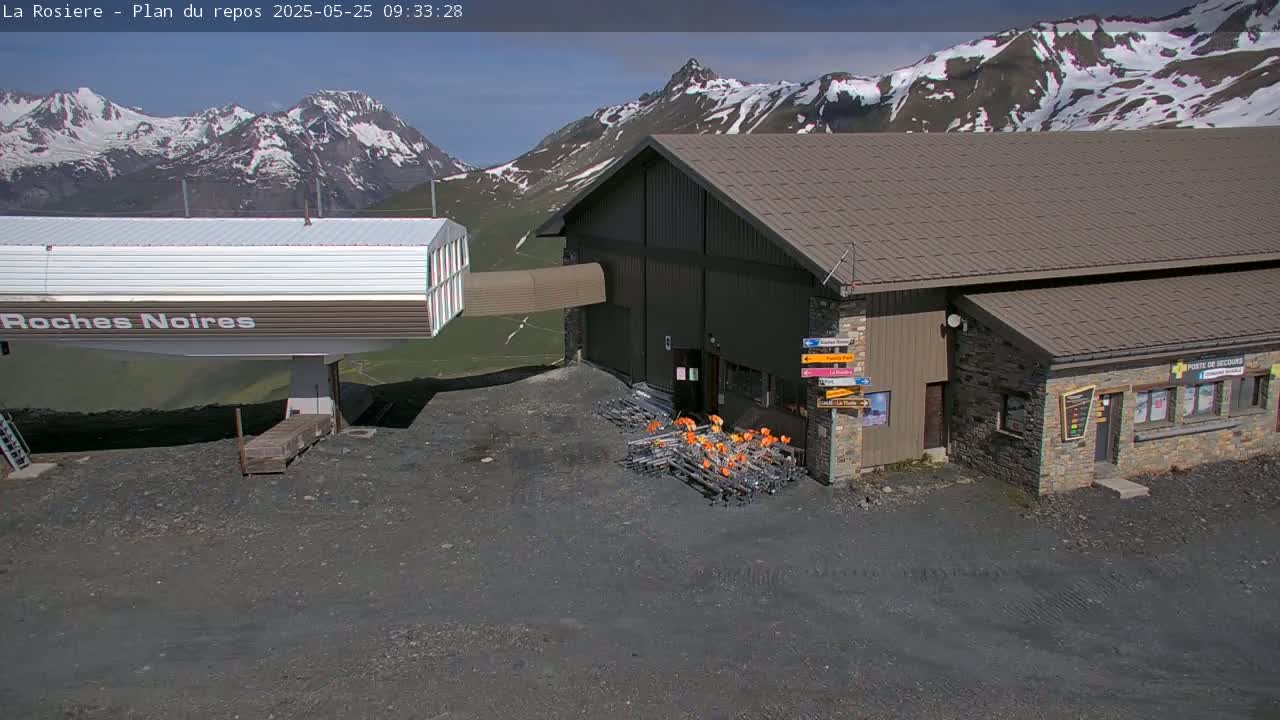 A ski lift station with a brown building sits in front of a mountain range with snow-capped peaks under a sunny sky.