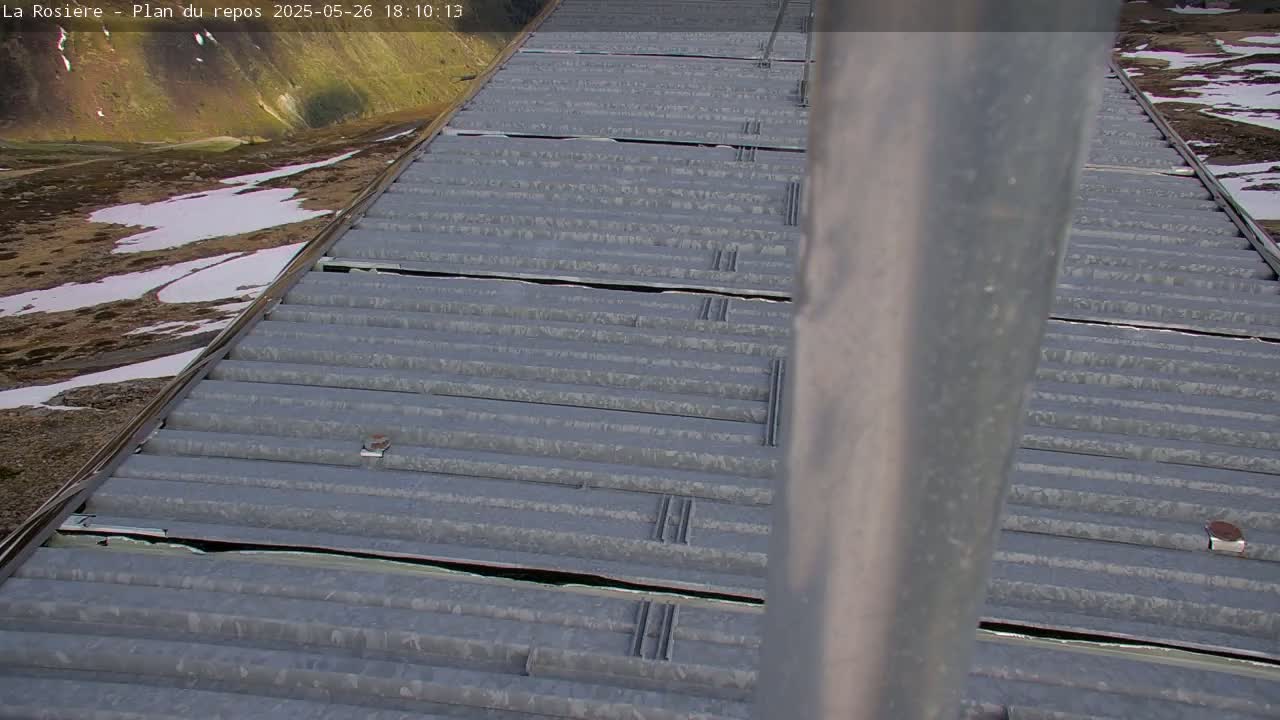 A corrugated metal surface with some damage is seen angled towards a mountainous landscape with patches of snow under partly sunny conditions.