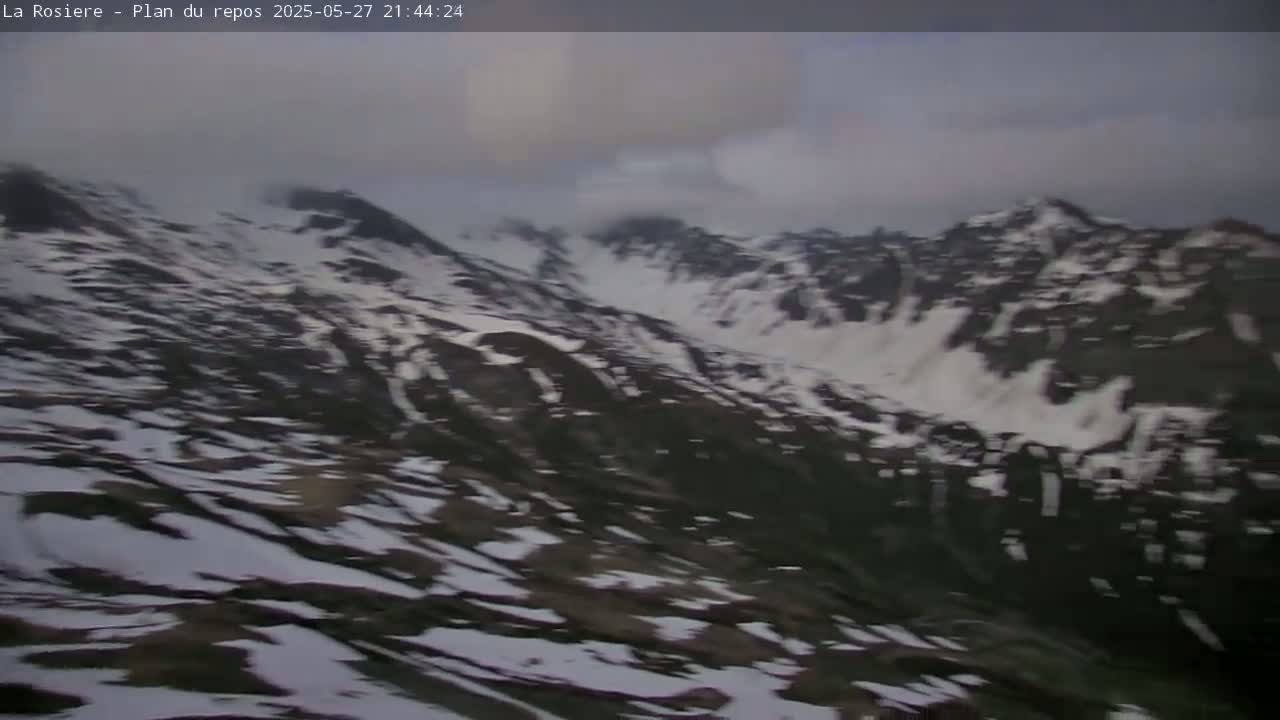 A high-angle view shows snow-dusted dark mountains under an overcast sky.
