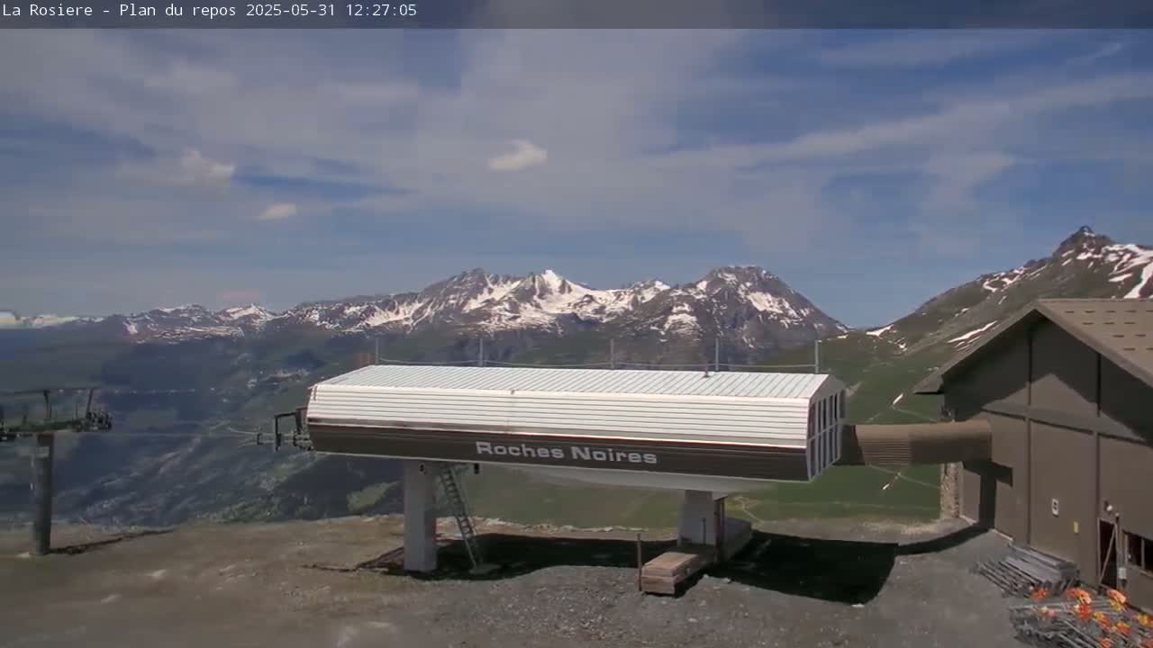 A ski lift station sits in the foreground, with snow-capped mountains and a partly cloudy sky visible in the background.