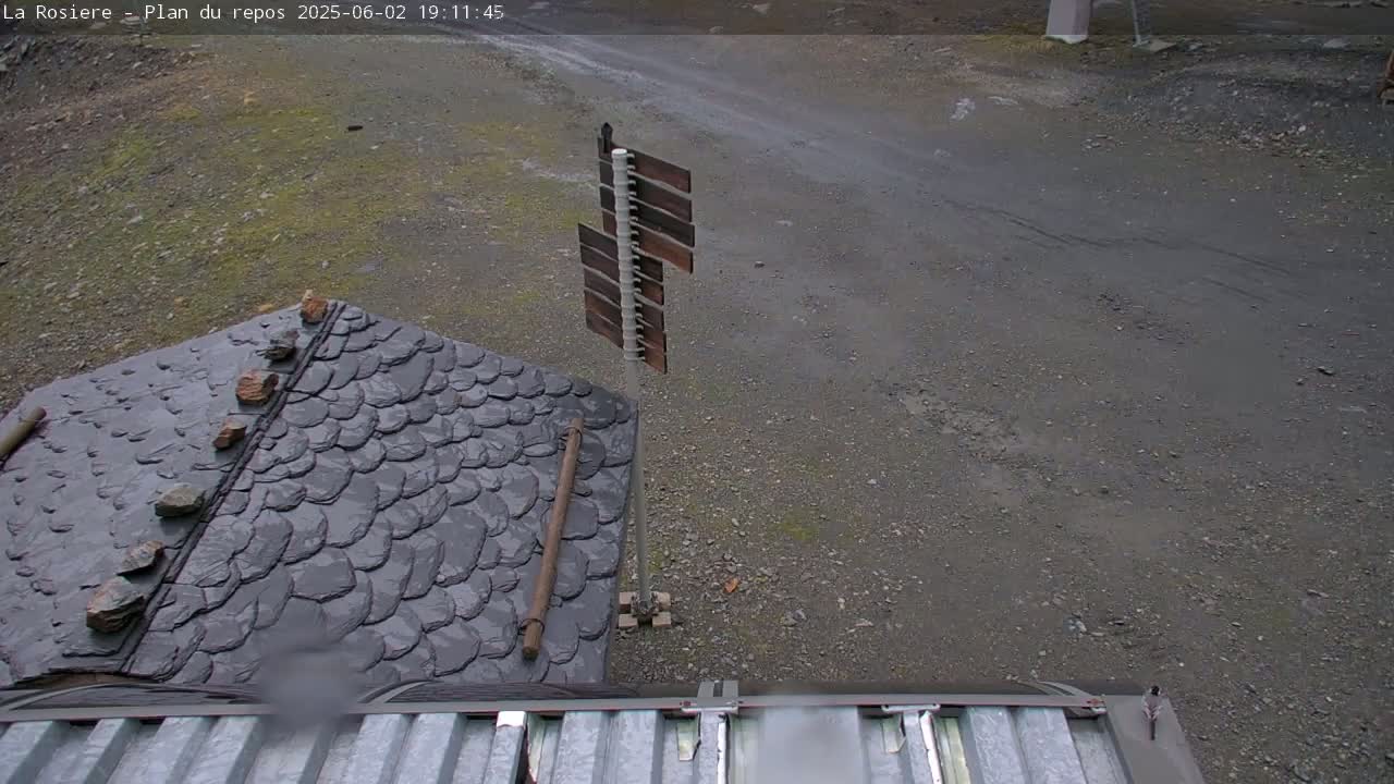 A dark gray, slate-tiled roof and a gravel area are visible, with a tall, thin wooden signpost in the background, under overcast skies.