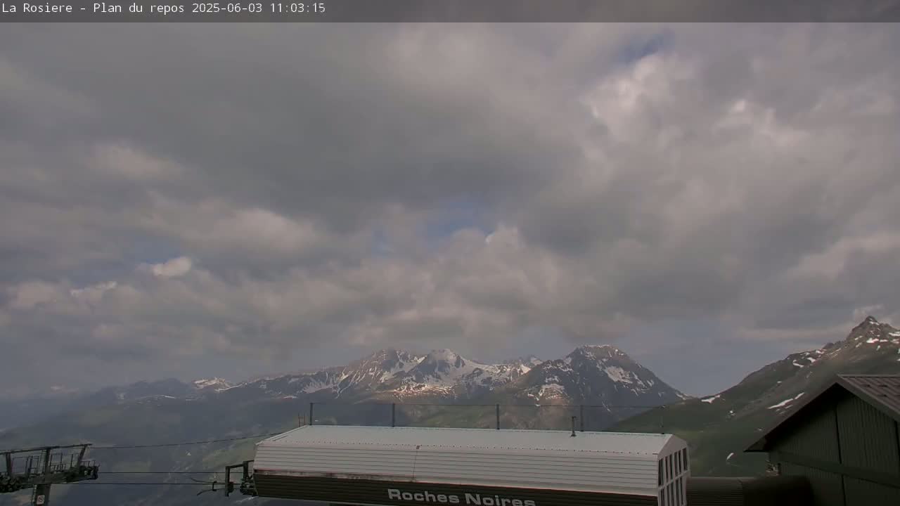 A mostly cloudy sky hangs over snow-capped mountains, partially visible behind a building and ski lift.