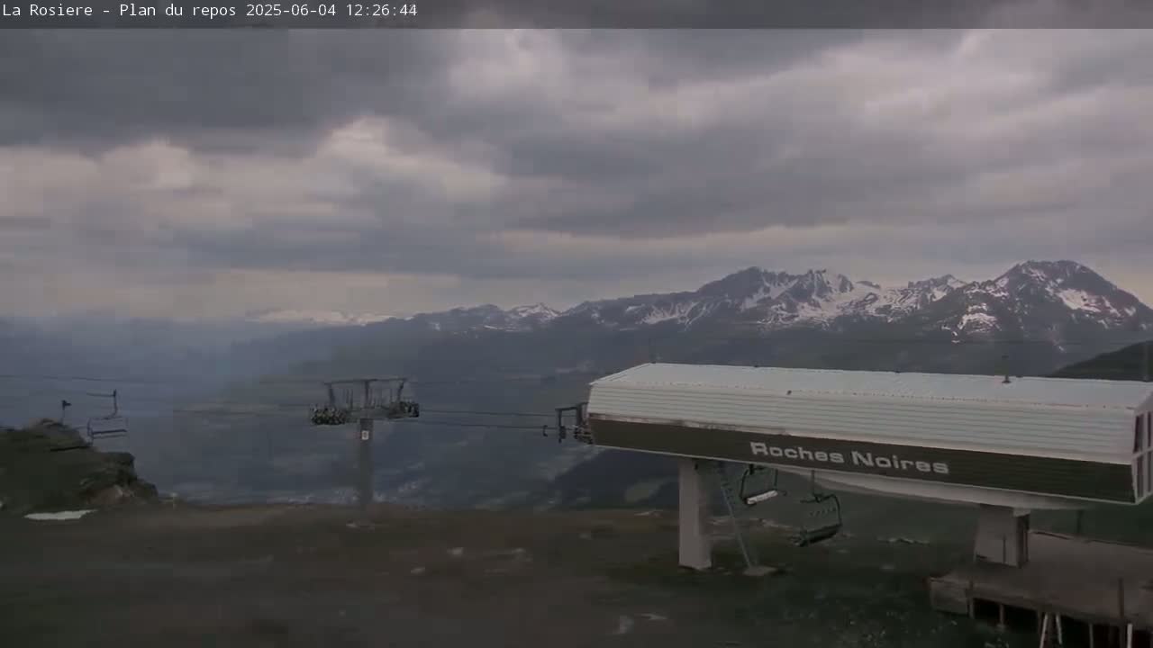 A ski lift station sits in the foreground, overlooking a valley and snow-capped mountains under a cloudy sky.