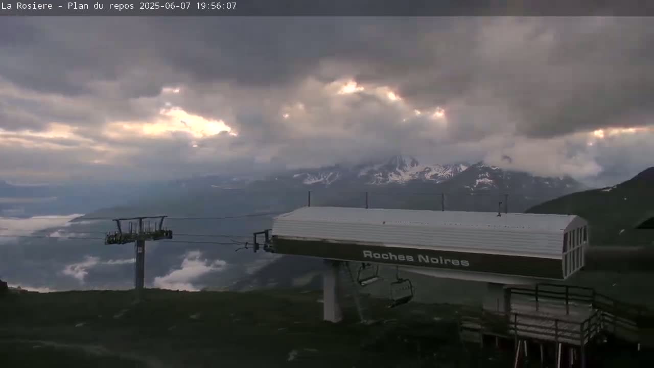 A chairlift station sits atop a mountain overlooking a valley shrouded in clouds under a partly cloudy sky.