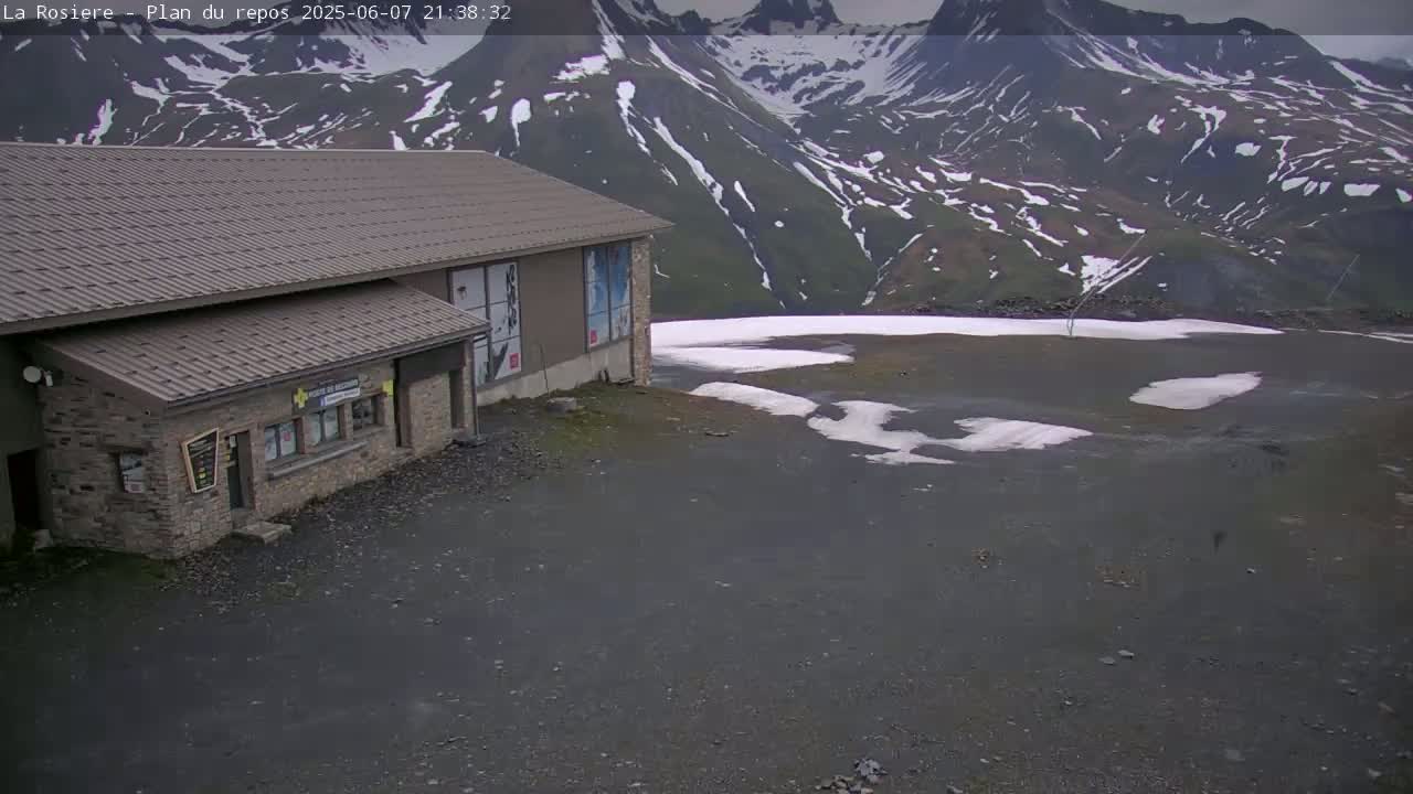 A stone building sits in a mountain area with patches of snow on the ground under an overcast sky.