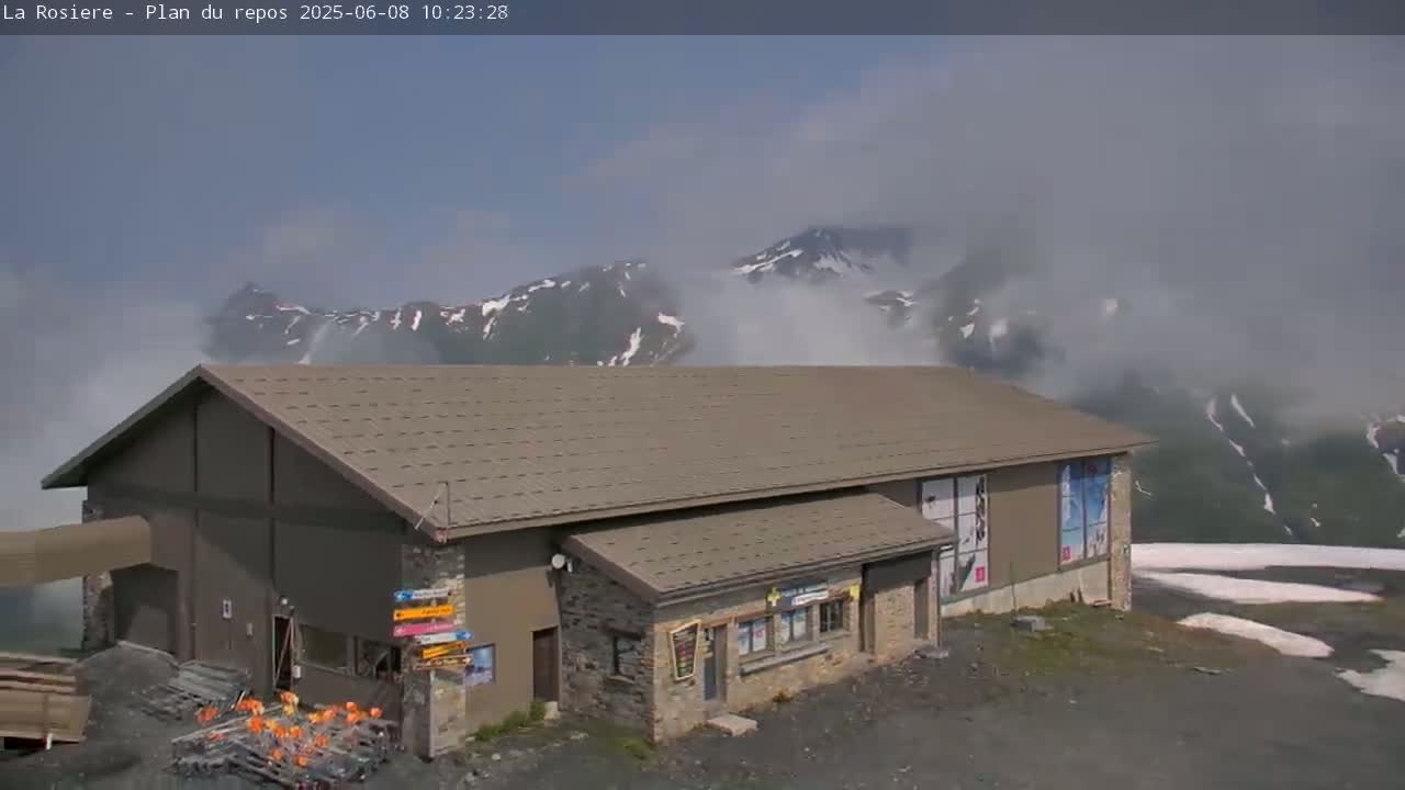 A brown, single-story building with a gabled roof sits on a mountainside partially obscured by fog, with snow visible on the distant peaks.