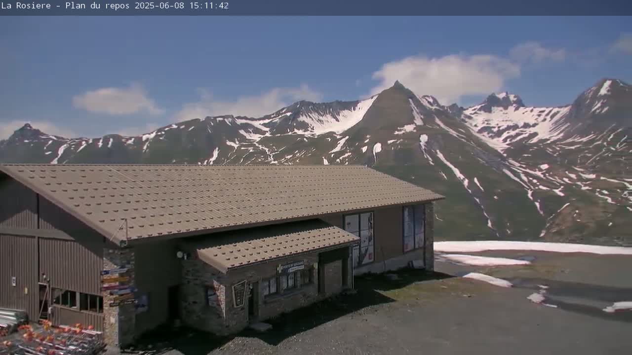 A brown building with a metal roof sits in front of a mountain range with patches of snow under a mostly clear, bright blue sky.
