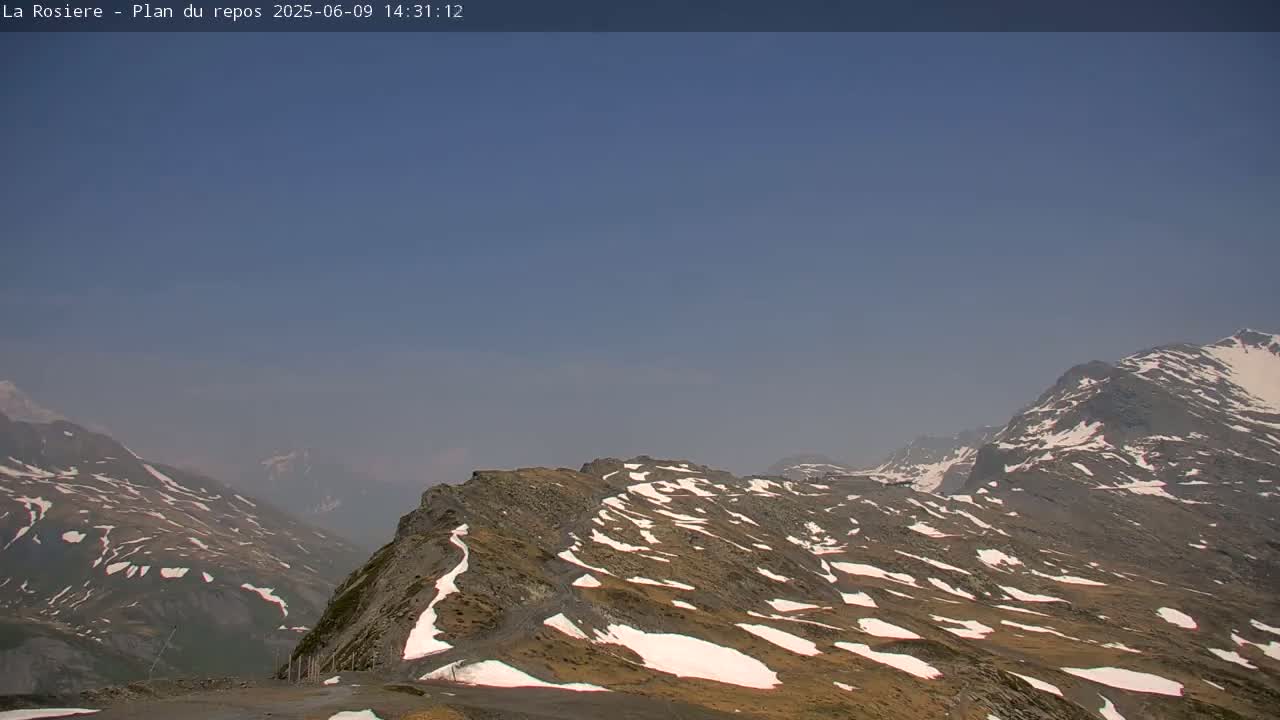 A hazy, clear-sky day reveals a mountain range with patches of snow on the brown and gray rock faces.
