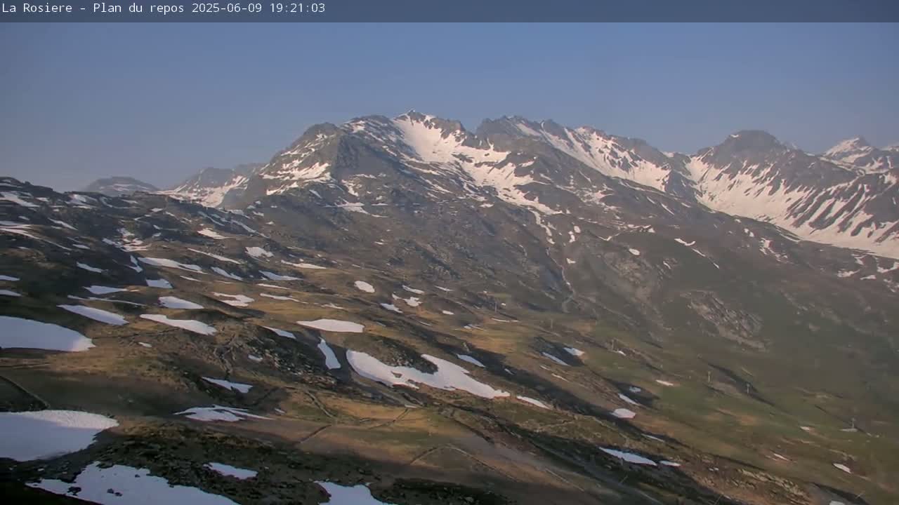A high-angle view shows a mountainous landscape with patches of snow under a clear, bright sky.