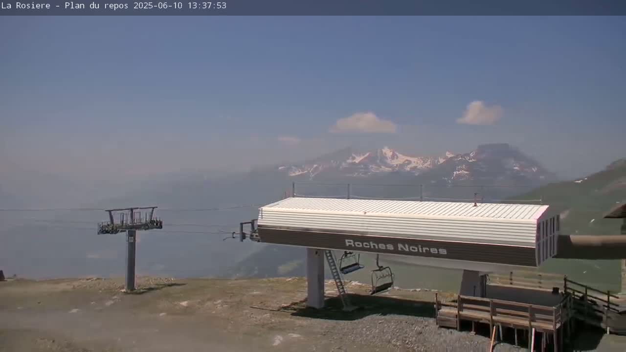 A ski lift station sits atop a mountain under a mostly clear, hazy sky, with snow-capped mountains visible in the distance.