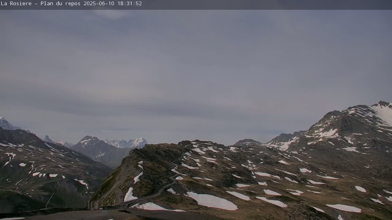 A winding road is visible on a mountain range with patches of snow under an overcast sky.