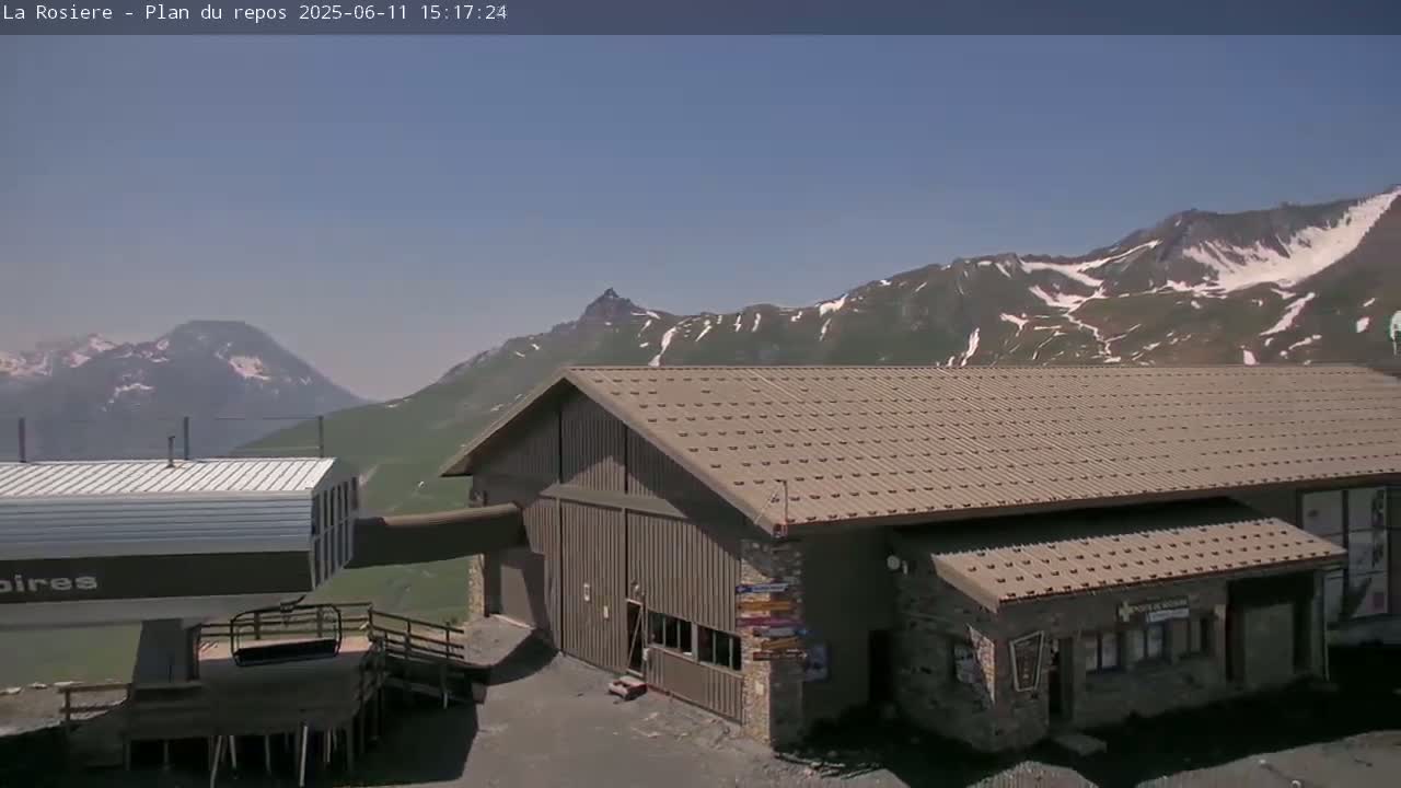 A brown building with a metal roof sits below snow-capped mountains on a clear, sunny day.