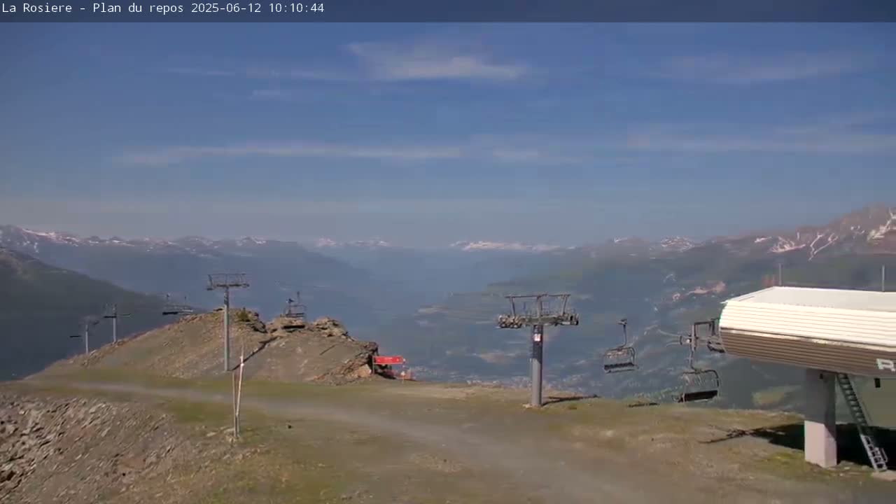 A mostly clear, sunny day overlooking a mountain valley with several ski lifts and a lift station visible in the foreground.