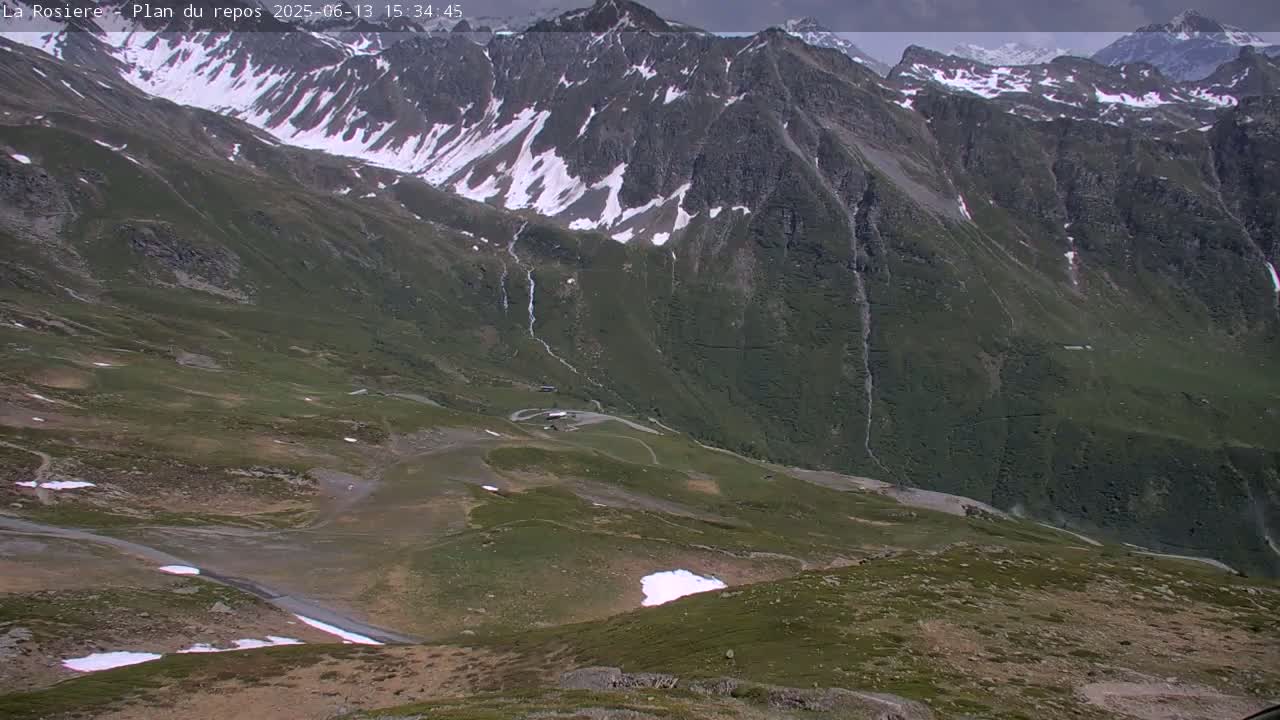 A high-angle view of a mountainous landscape under partly cloudy skies shows a winding road and patches of snow amidst green hillsides and rocky mountain peaks.