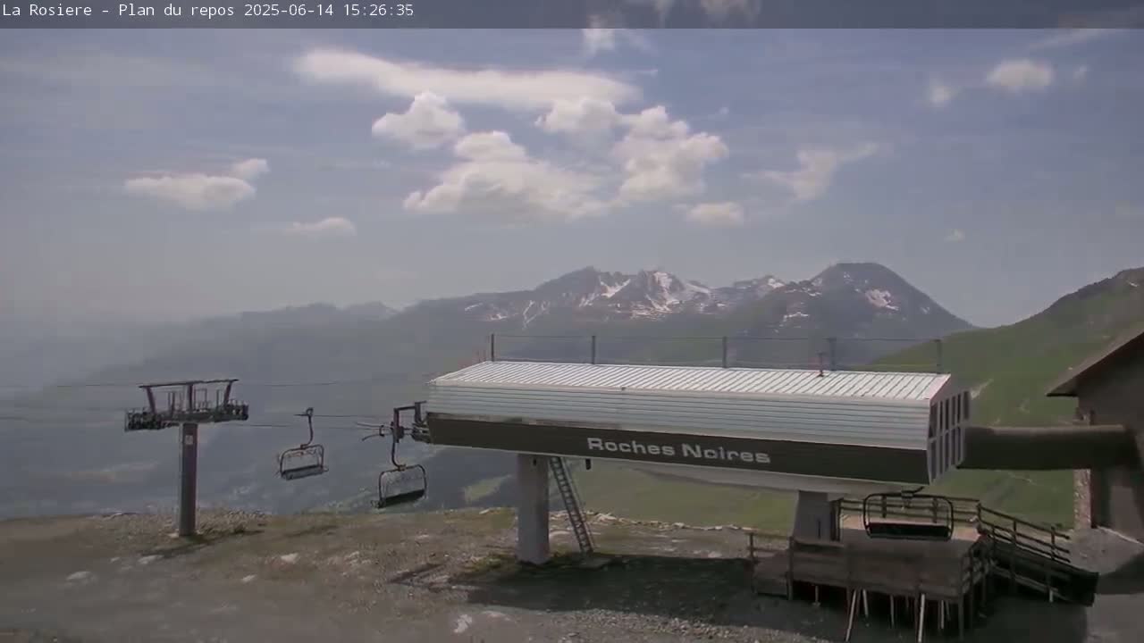 A chairlift station sits atop a mountain, overlooking a partly cloudy sky and snow-capped mountains in the distance.