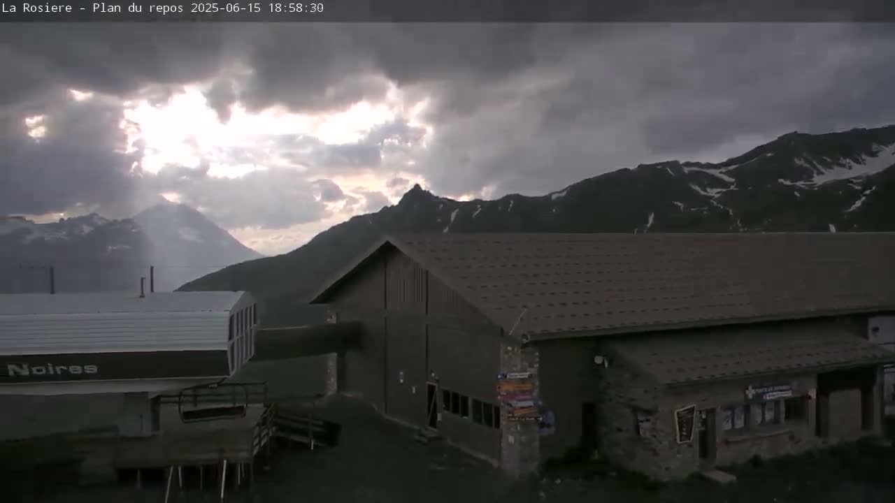A dark-colored building sits below a cloudy sky, partially illuminated by sunlight breaking through the clouds behind snow-capped mountains.