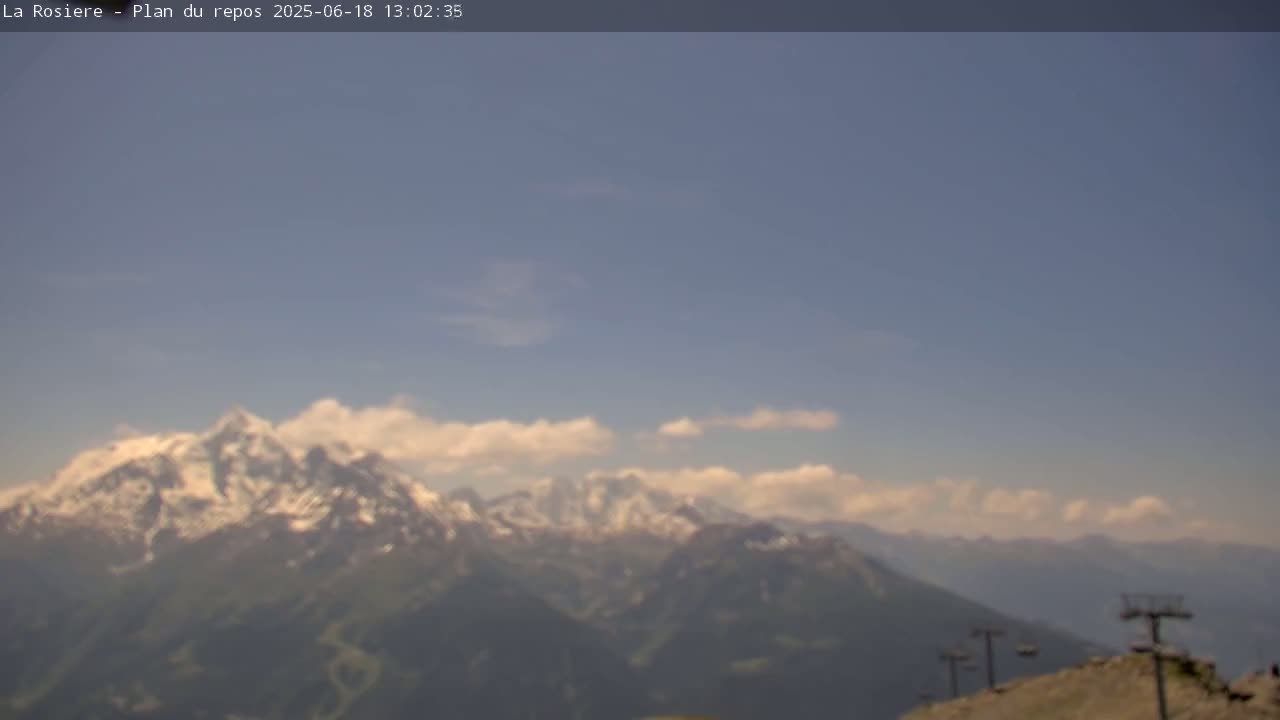 A mostly clear, bright sky overlooks snow-capped mountains and a few chairlifts on a hilltop in the foreground.