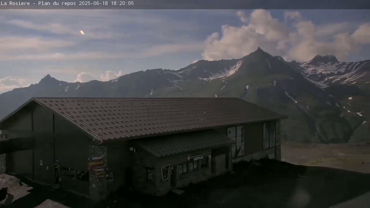 A dark-colored building with a sloped roof sits in front of snow-capped mountains under a partly cloudy sky.