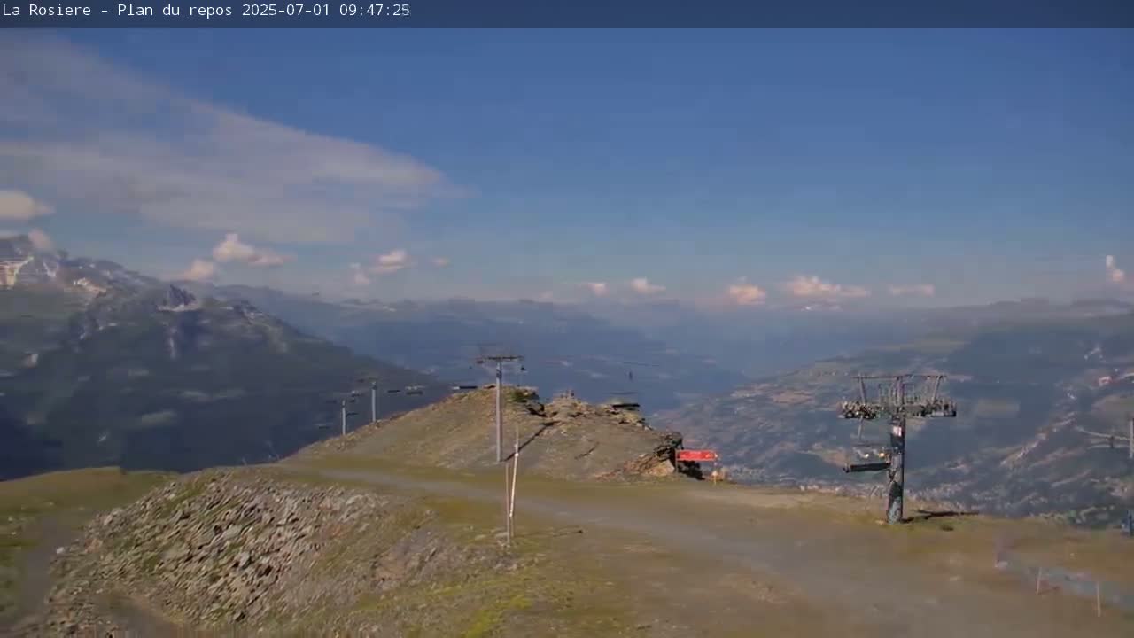 A mostly sunny day reveals a mountaintop view of a valley with chairlifts and a rocky, sparsely vegetated landscape.