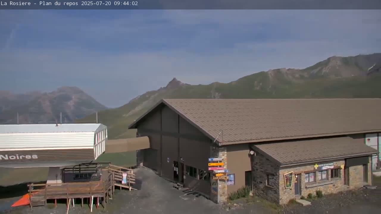A ski lift station building sits below a mountain range on a mostly sunny day.