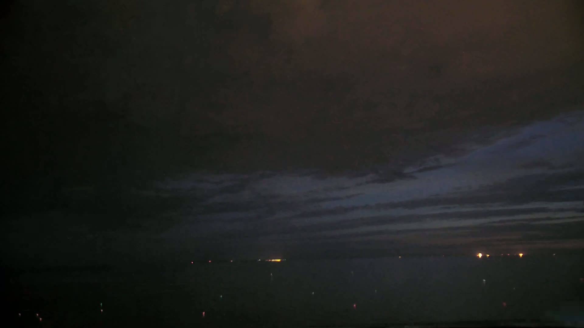 A dark, cloudy night sky over a body of water with a few distant lights visible on the horizon.