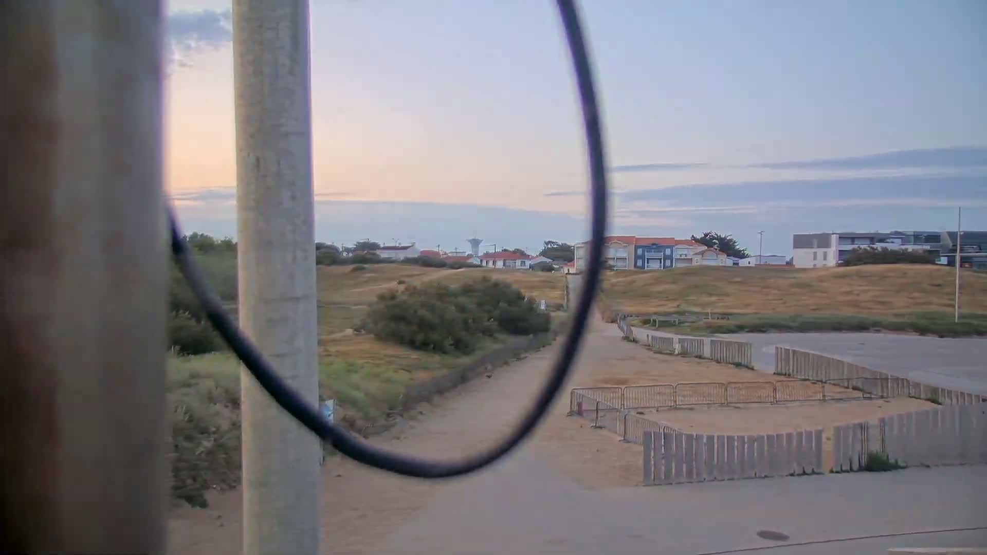 A view through a looped cable shows a sandy area with a wooden fence, a path, and houses in the distance under a pale, clear sky.