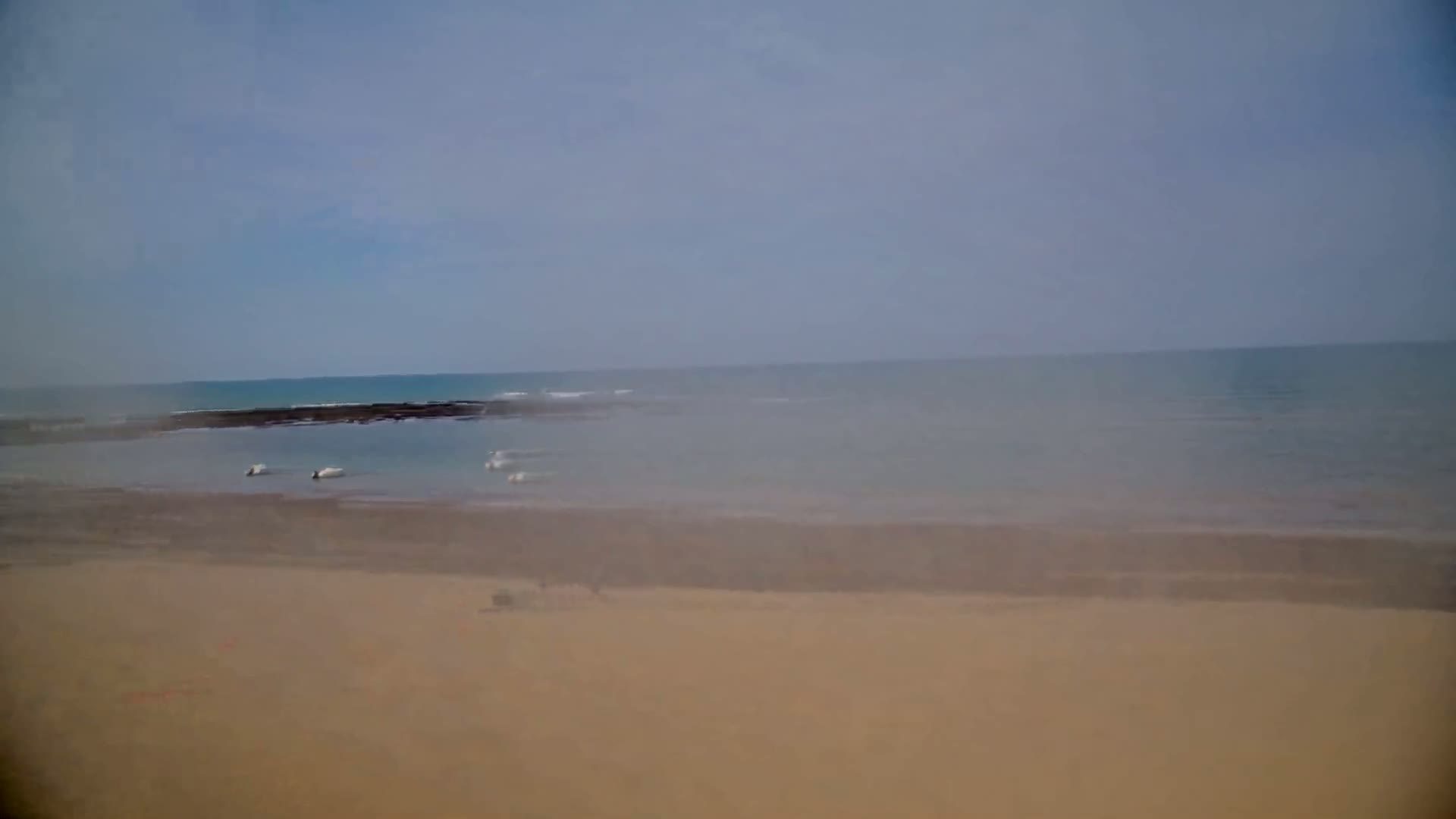 A hazy, sandy beach stretches to a calm ocean with dark rocks visible near the shore.