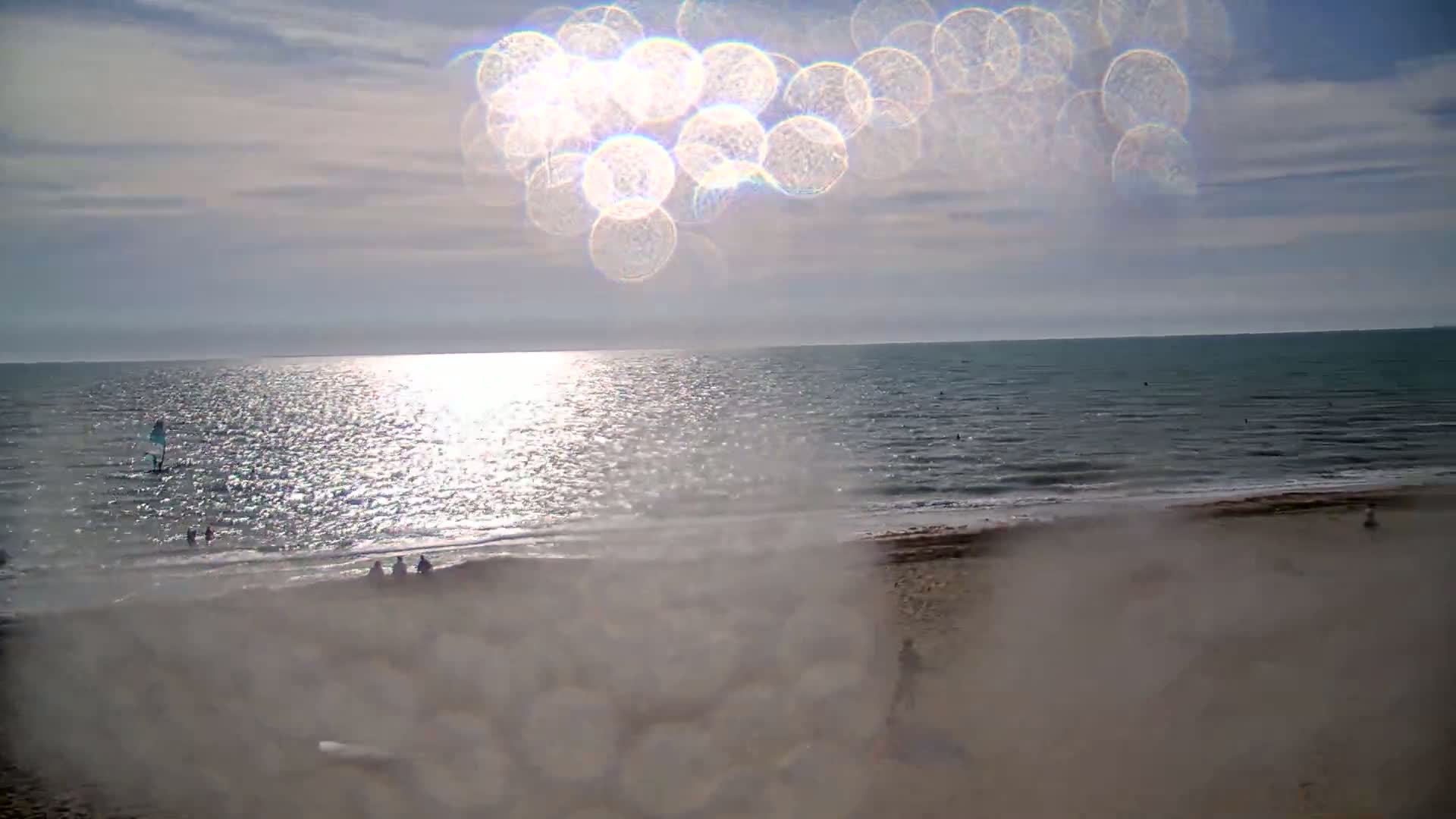 A sunny day at a beach shows a calm sea with a few people in the water and on the sand, viewed through a lens with a significant glare.