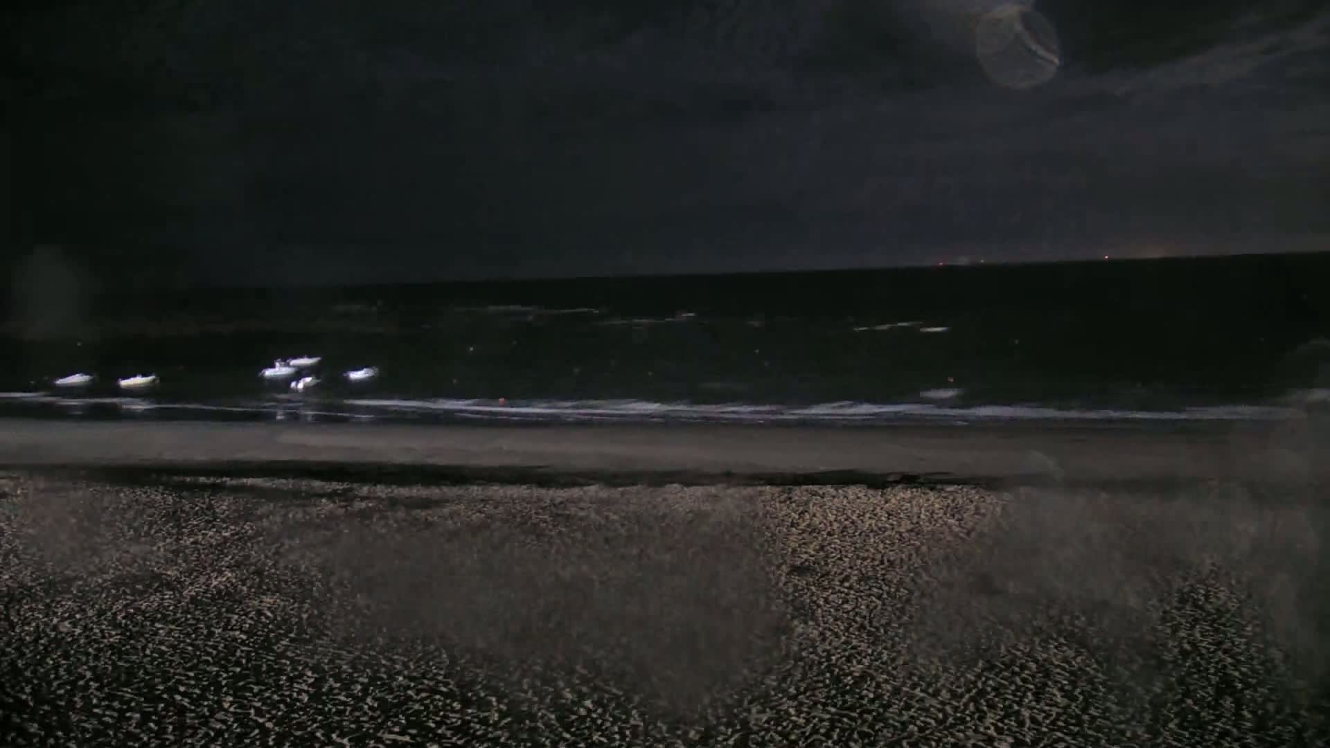 A dark, nighttime beach scene shows several small boats on a calm, dark ocean with gentle waves lapping a sandy shore.