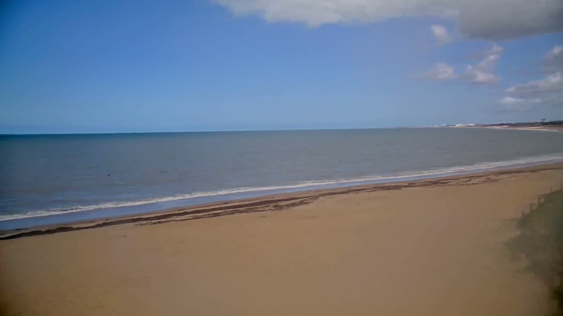 A wide sandy beach meets a calm, gray-blue ocean under a mostly sunny sky with scattered clouds.