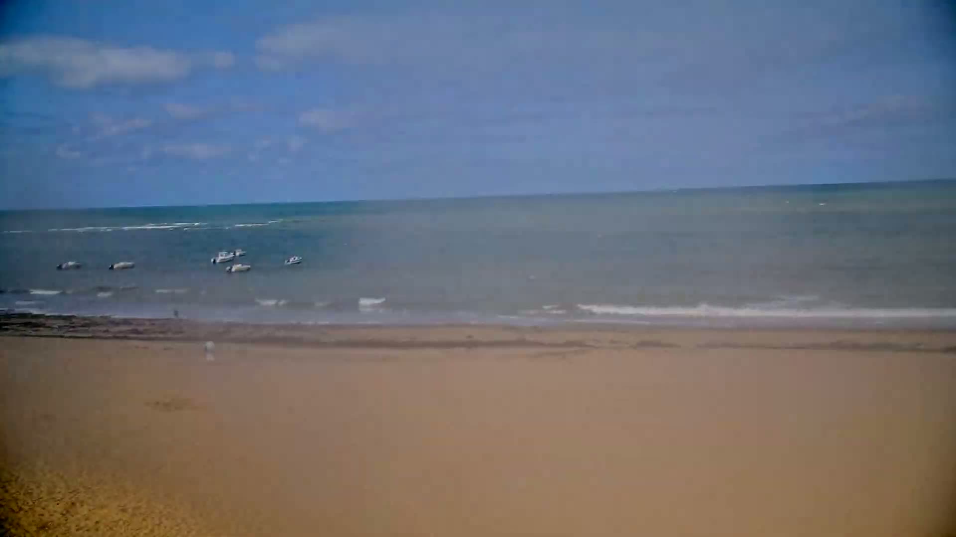 A sandy beach stretches to a calm sea where several small boats are visible under a partly cloudy sky.