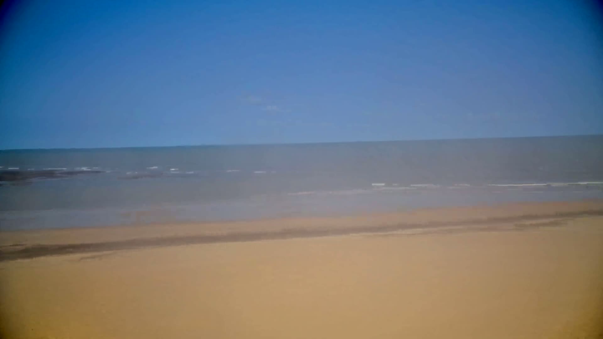 A hazy, light-brown sand beach meets a calm, gray-blue ocean under a clear blue sky.