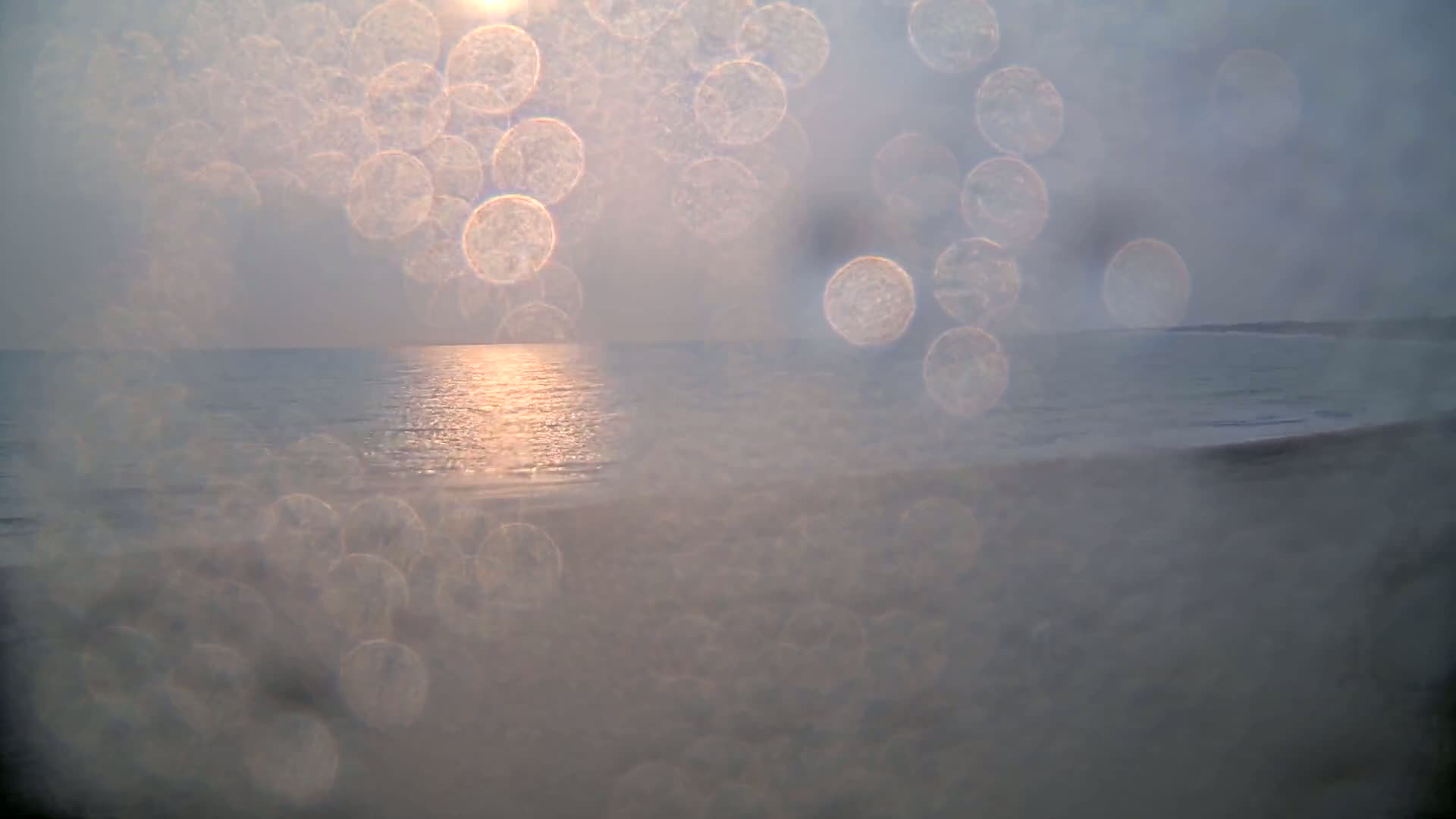 A blurry, sunlit ocean scene is viewed through a wet surface, with many water droplets creating bokeh effects.