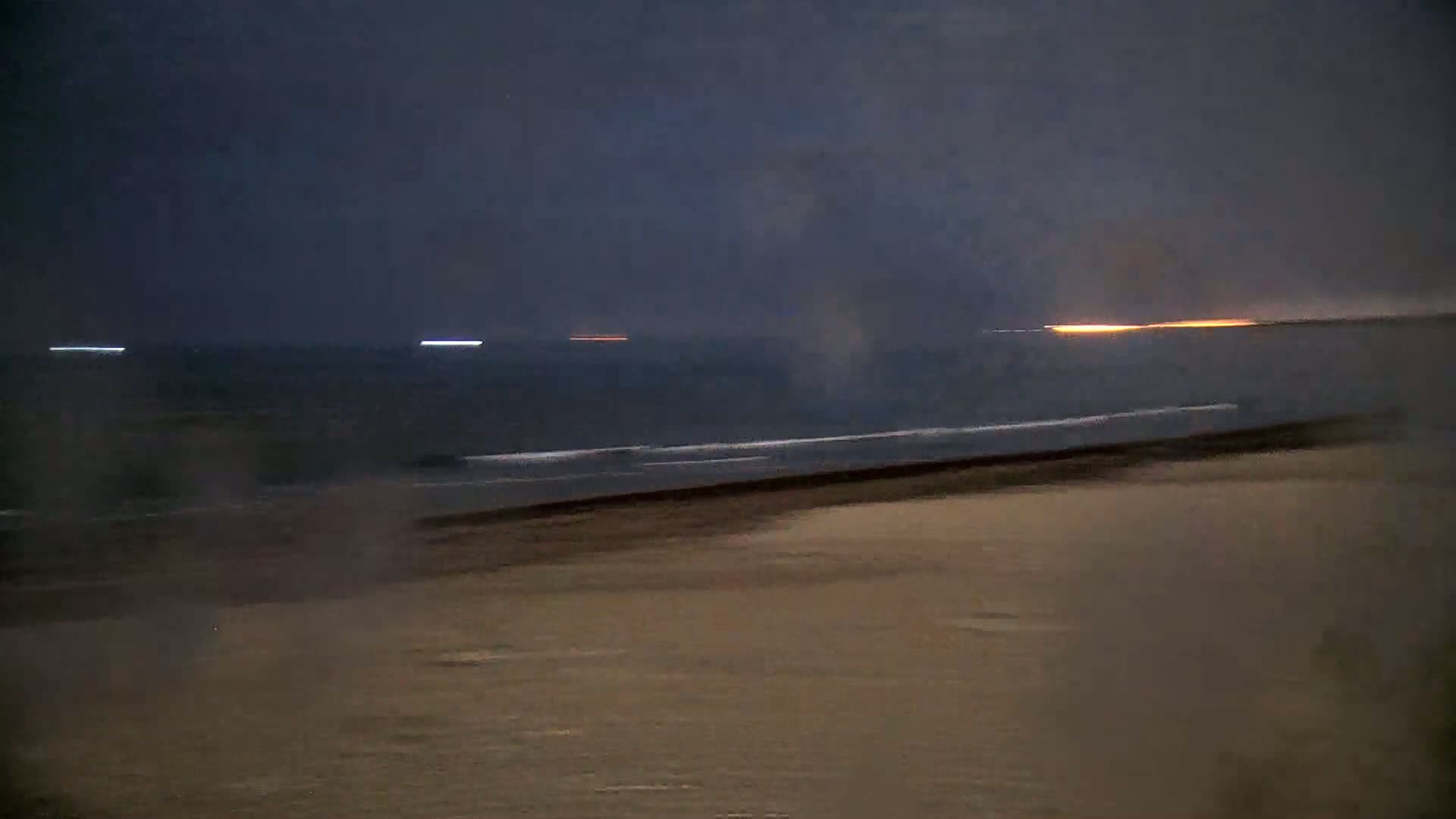 A dark beach at night with a calm ocean and faint lights on the horizon.